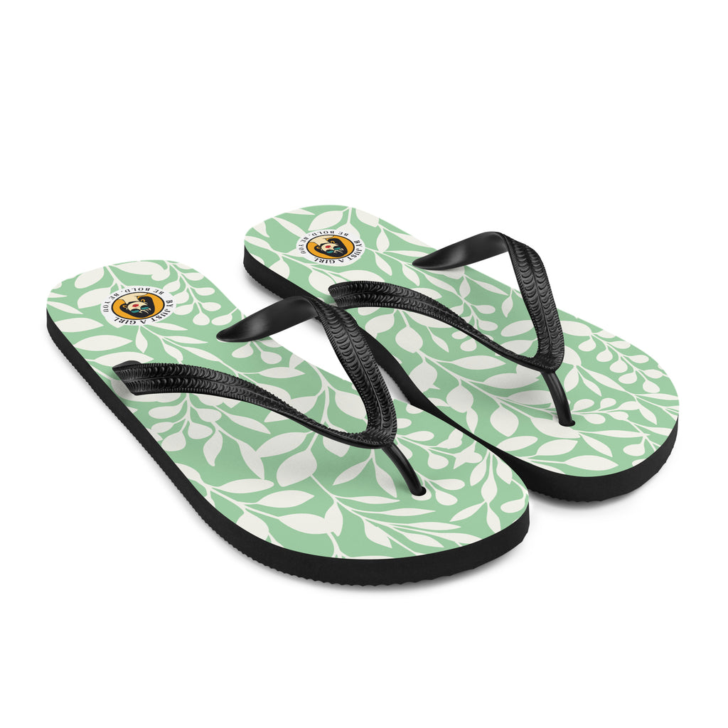 Ein Paar Flip-Flops mit einem hellmintgrünen Untergrund, der mit einem wiederkehrenden Muster aus stilisierten weißen Blättern und kleinen runden Beeren bedeckt ist. Die Riemen sind schwarz und haben eine strukturierte Oberfläche. Auf der Ferse jedes Flip-Flops befindet sich ein rundes gelbes Emblem mit dem Text "BY JUST A GIRL" und "BE BOLD. BE YOU" sowie dem stilisierten Kopf einer Frau mit Bob-Frisur und Sonnenbrille.