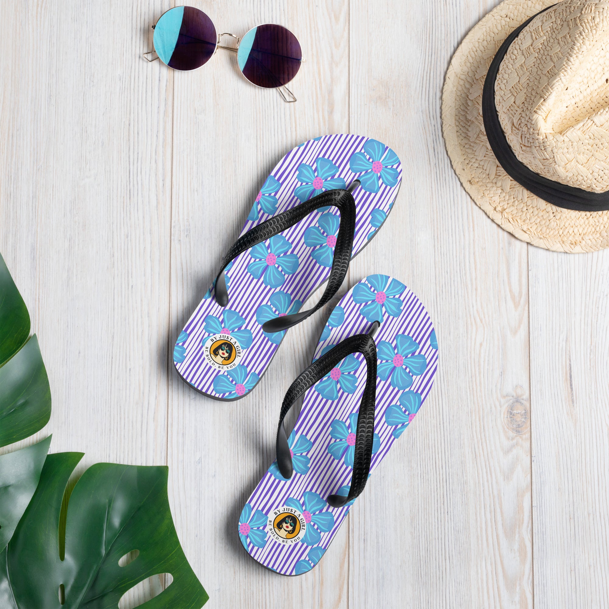 Ein Paar Flip-Flops mit einem Obermaterial, das ein Muster aus vertikalen lila und weißen Streifen zeigt, überlagert von großen, stilisierten blauen Blüten mit pinkfarbenen Zentren. Die Riemen sind schwarz und haben eine strukturierte Oberfläche. Auf der Ferse jedes Flip-Flops befindet sich ein rundes gelbes Emblem mit dem Text "BY JUST A GIRL" und "BE BOLD. BE YOU" sowie dem stilisierten Kopf einer Frau mit Bob-Frisur und Sonnenbrille.