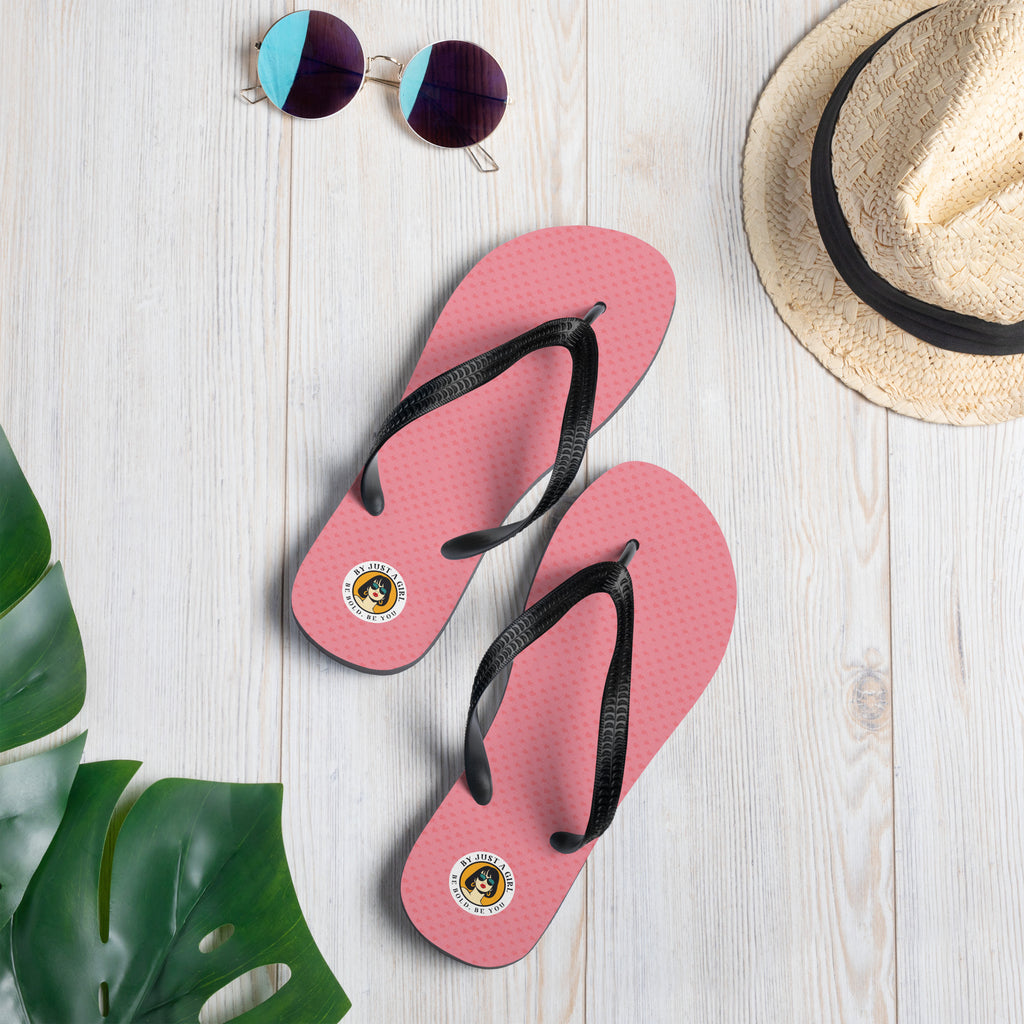 Ein Paar Flip-Flops mit einem Obermaterial, kleine Herzen in Rosa und Rot zeigt. Die Riemen sind schwarz und haben eine strukturierte Oberfläche. Auf der Ferse jedes Flip-Flops befindet sich ein rundes gelbes Emblem mit dem Text "BY JUST A GIRL" und "BE BOLD. BE YOU" sowie dem stilisierten Kopf einer Frau mit Bob-Frisur und Sonnenbrille.