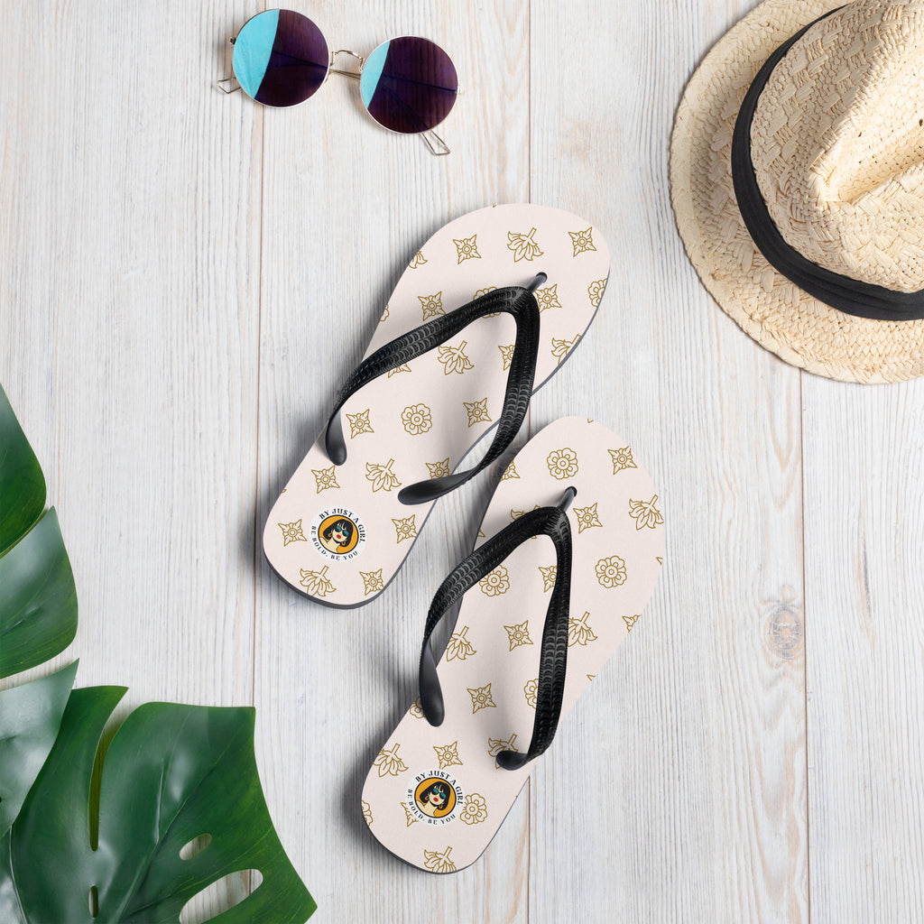 Ein Paar Flip-Flops mit einem hellbeigen oder cremefarbenen Obermaterial, das mit einem wiederkehrenden Muster aus stilisierten goldenen Lotusblüten und anderen floralen oder blattartigen Ornamenten bedeckt ist. Die Riemen sind schwarz und haben eine strukturierte Oberfläche. Auf der Ferse jedes Flip-Flops befindet sich ein rundes gelbes Emblem mit dem Text "BY JUST A GIRL" und "BE BOLD. BE YOU" sowie dem stilisierten Kopf einer Frau mit Bob-Frisur und Sonnenbrille.
