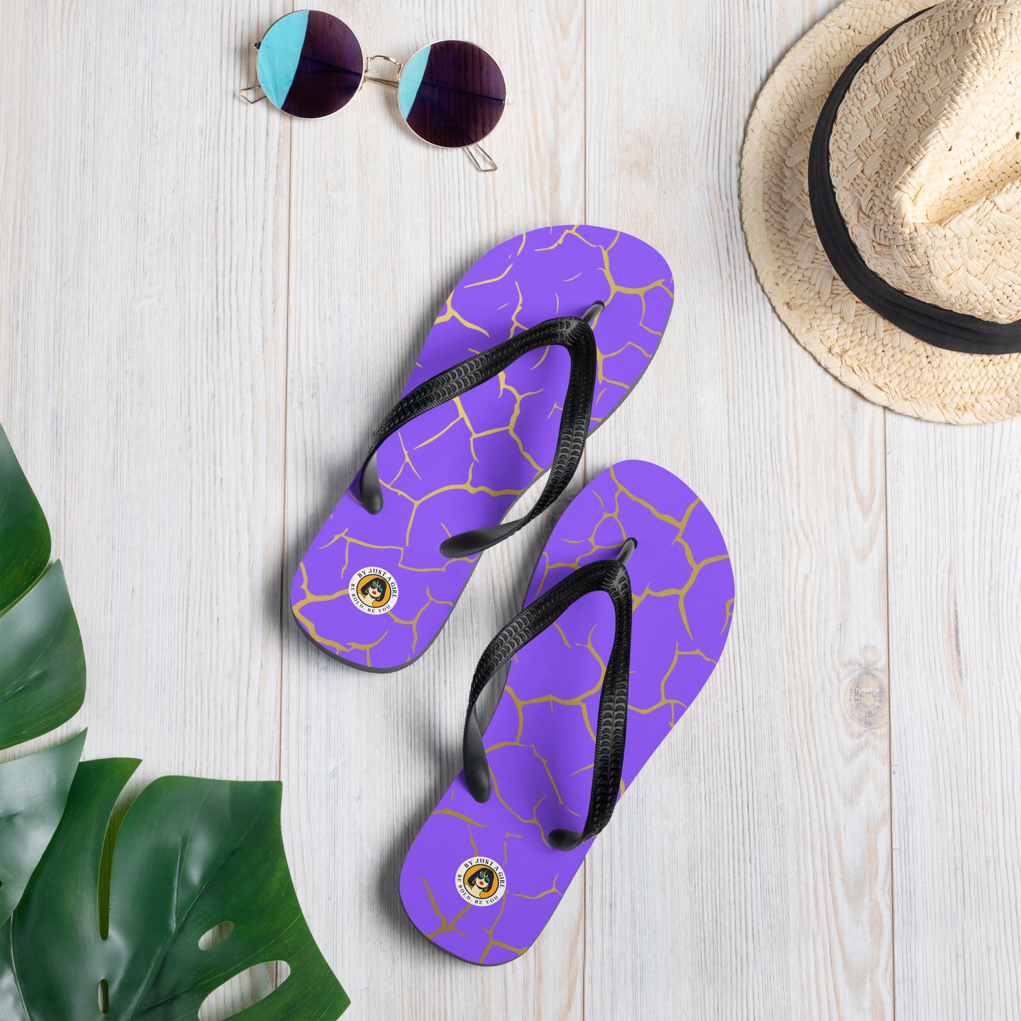 Ein Paar Flip-Flops mit einem Obermaterial in einem kräftigen Violett, das von einem unregelmäßigen, spinnennetzartigen Muster aus goldenen Linien durchzogen ist, die wie Risse aussehen. Die Riemen sind schwarz und haben eine strukturierte Oberfläche. Auf der Ferse jedes Flip-Flops befindet sich ein rundes gelbes Emblem mit dem Text "BY JUST A GIRL" und "BE BOLD. BE YOU" sowie dem stilisierten Kopf einer Frau mit Bob-Frisur und Sonnenbrille.