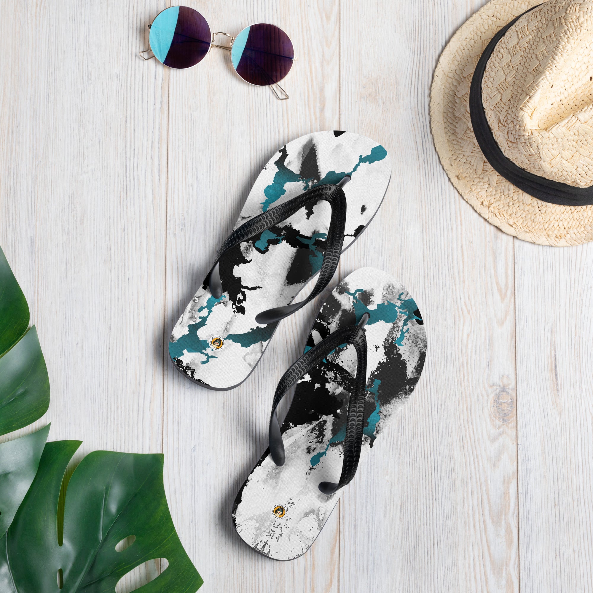 Ein Paar schwarze Herren-Flip-Flops mit einem Obermaterial, das ein abstraktes, dynamisches Muster aus unregelmäßigen schwarzen, weißen und türkisen Farbflächen oder Spritzern auf einem hellen Hintergrund zeigt. Die Riemen sind schwarz und haben eine strukturierte Oberfläche.