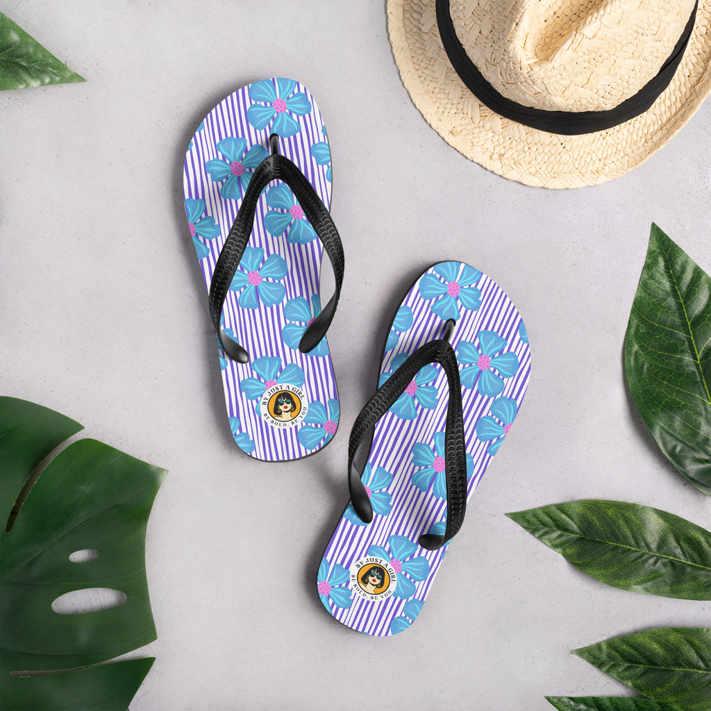 Ein Paar Flip-Flops mit einem Obermaterial, das ein Muster aus vertikalen lila und weißen Streifen zeigt, überlagert von großen, stilisierten blauen Blüten mit pinkfarbenen Zentren. Die Riemen sind schwarz und haben eine strukturierte Oberfläche. Auf der Ferse jedes Flip-Flops befindet sich ein rundes gelbes Emblem mit dem Text "BY JUST A GIRL" und "BE BOLD. BE YOU" sowie dem stilisierten Kopf einer Frau mit Bob-Frisur und Sonnenbrille.