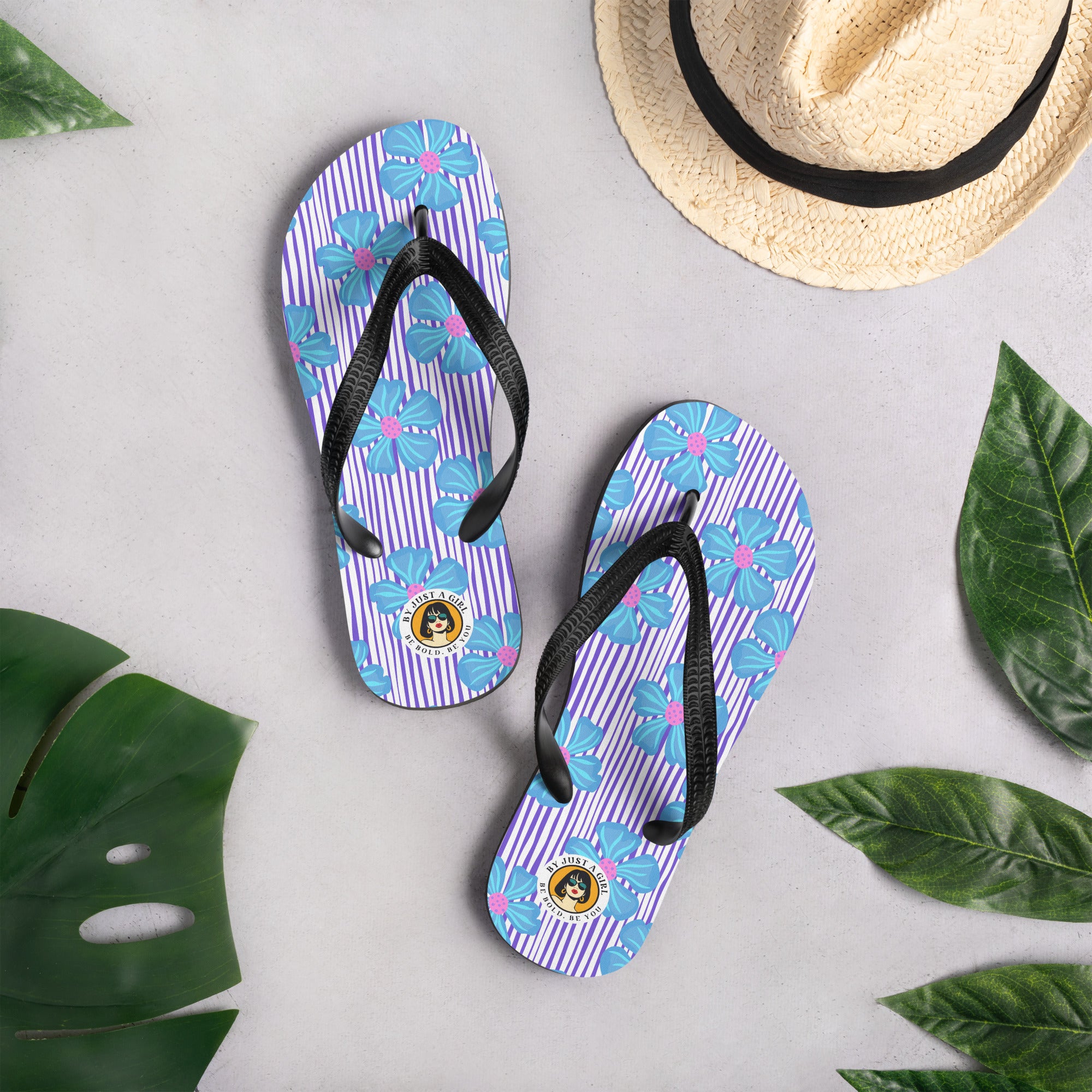 Ein Paar Flip-Flops mit einem Obermaterial, das ein Muster aus vertikalen lila und weißen Streifen zeigt, überlagert von großen, stilisierten blauen Blüten mit pinkfarbenen Zentren. Die Riemen sind schwarz und haben eine strukturierte Oberfläche. Auf der Ferse jedes Flip-Flops befindet sich ein rundes gelbes Emblem mit dem Text "BY JUST A GIRL" und "BE BOLD. BE YOU" sowie dem stilisierten Kopf einer Frau mit Bob-Frisur und Sonnenbrille.