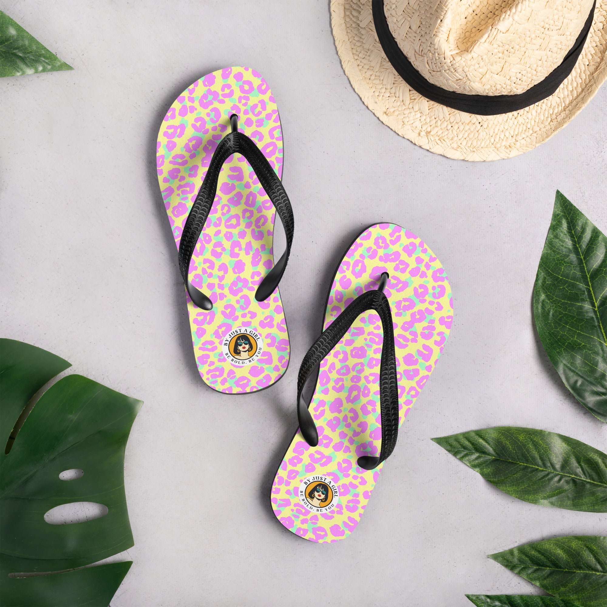 Ein Paar Flip-Flops mit einem Obermaterial, das ein abstraktes Leopardenmuster in ungewöhnlichen, leuchtenden Farben zeigt: Pinke und mintgrüne Flecken auf einem hellgelben oder zitronengelben Hintergrund. Die Riemen sind schwarz und haben eine strukturierte Oberfläche. Auf der Ferse jedes Flip-Flops befindet sich ein rundes gelbes Emblem mit dem Text "BY JUST A GIRL" und "BE BOLD. BE YOU" sowie dem stilisierten Kopf einer Frau mit Bob-Frisur und Sonnenbrille.