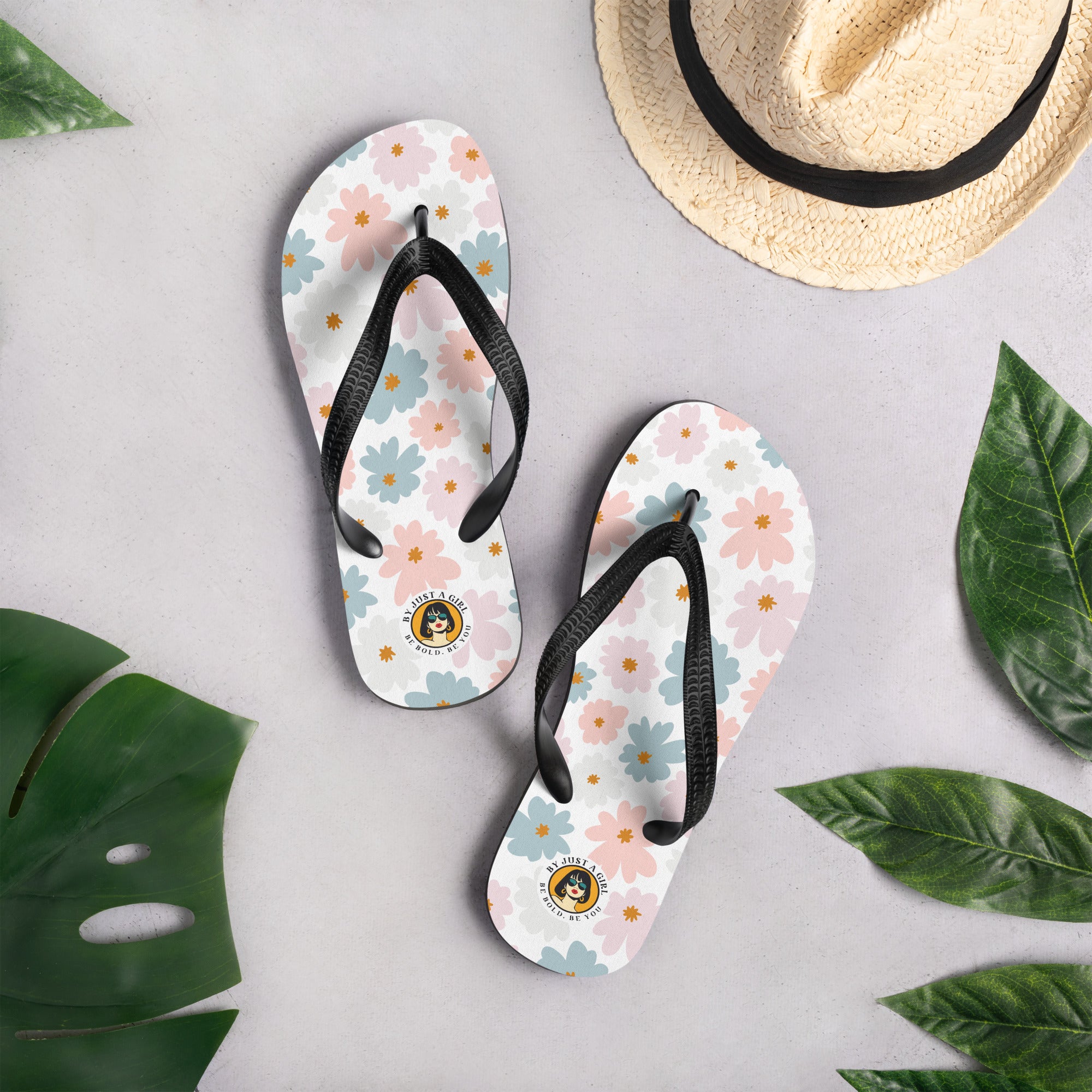 Ein Paar Flip-Flops mit einem weißen Obermaterial, das mit einem wiederkehrenden Muster aus stilisierten, fünfblättrigen Blüten in Pastellfarben wie Hellrosa, Hellblau und Beige bedeckt ist, ergänzt durch kleine orangefarbene Sterne in der Mitte einiger Blüten. Die Riemen sind schwarz und haben eine strukturierte Oberfläche. Auf der Ferse jedes Flip-Flops befindet sich ein rundes gelbes Emblem mit dem Text "BY JUST A GIRL" und "BE BOLD. BE YOU" sowie dem stilisierten Kopf einer Frau mit Bob-Frisur und Sonne