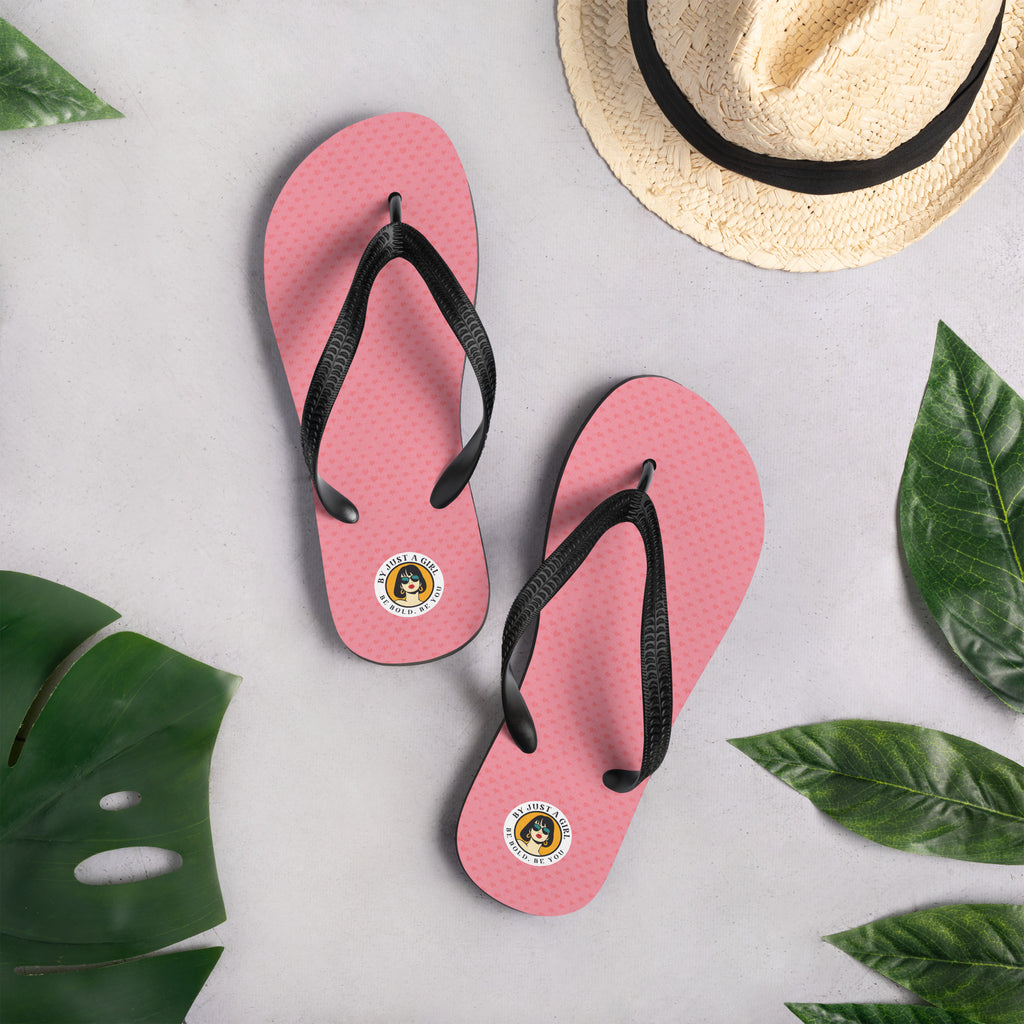 Ein Paar Flip-Flops mit einem Obermaterial, kleine Herzen in Rosa und Rot zeigt. Die Riemen sind schwarz und haben eine strukturierte Oberfläche. Auf der Ferse jedes Flip-Flops befindet sich ein rundes gelbes Emblem mit dem Text "BY JUST A GIRL" und "BE BOLD. BE YOU" sowie dem stilisierten Kopf einer Frau mit Bob-Frisur und Sonnenbrille.