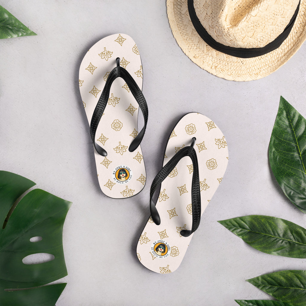 Ein Paar Flip-Flops mit einem hellbeigen oder cremefarbenen Obermaterial, das mit einem wiederkehrenden Muster aus stilisierten goldenen Lotusblüten und anderen floralen oder blattartigen Ornamenten bedeckt ist. Die Riemen sind schwarz und haben eine strukturierte Oberfläche. Auf der Ferse jedes Flip-Flops befindet sich ein rundes gelbes Emblem mit dem Text "BY JUST A GIRL" und "BE BOLD. BE YOU" sowie dem stilisierten Kopf einer Frau mit Bob-Frisur und Sonnenbrille.