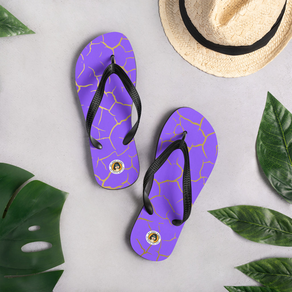 Ein Paar Flip-Flops mit einem Obermaterial in einem kräftigen Violett, das von einem unregelmäßigen, spinnennetzartigen Muster aus goldenen Linien durchzogen ist, die wie Risse aussehen. Die Riemen sind schwarz und haben eine strukturierte Oberfläche. Auf der Ferse jedes Flip-Flops befindet sich ein rundes gelbes Emblem mit dem Text "BY JUST A GIRL" und "BE BOLD. BE YOU" sowie dem stilisierten Kopf einer Frau mit Bob-Frisur und Sonnenbrille.