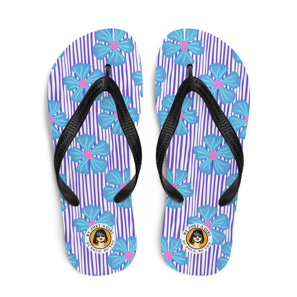 Ein Paar Flip-Flops mit einem Obermaterial, das ein Muster aus vertikalen lila und weißen Streifen zeigt, überlagert von großen, stilisierten blauen Blüten mit pinkfarbenen Zentren. Die Riemen sind schwarz und haben eine strukturierte Oberfläche. Auf der Ferse jedes Flip-Flops befindet sich ein rundes gelbes Emblem mit dem Text "BY JUST A GIRL" und "BE BOLD. BE YOU" sowie dem stilisierten Kopf einer Frau mit Bob-Frisur und Sonnenbrille.