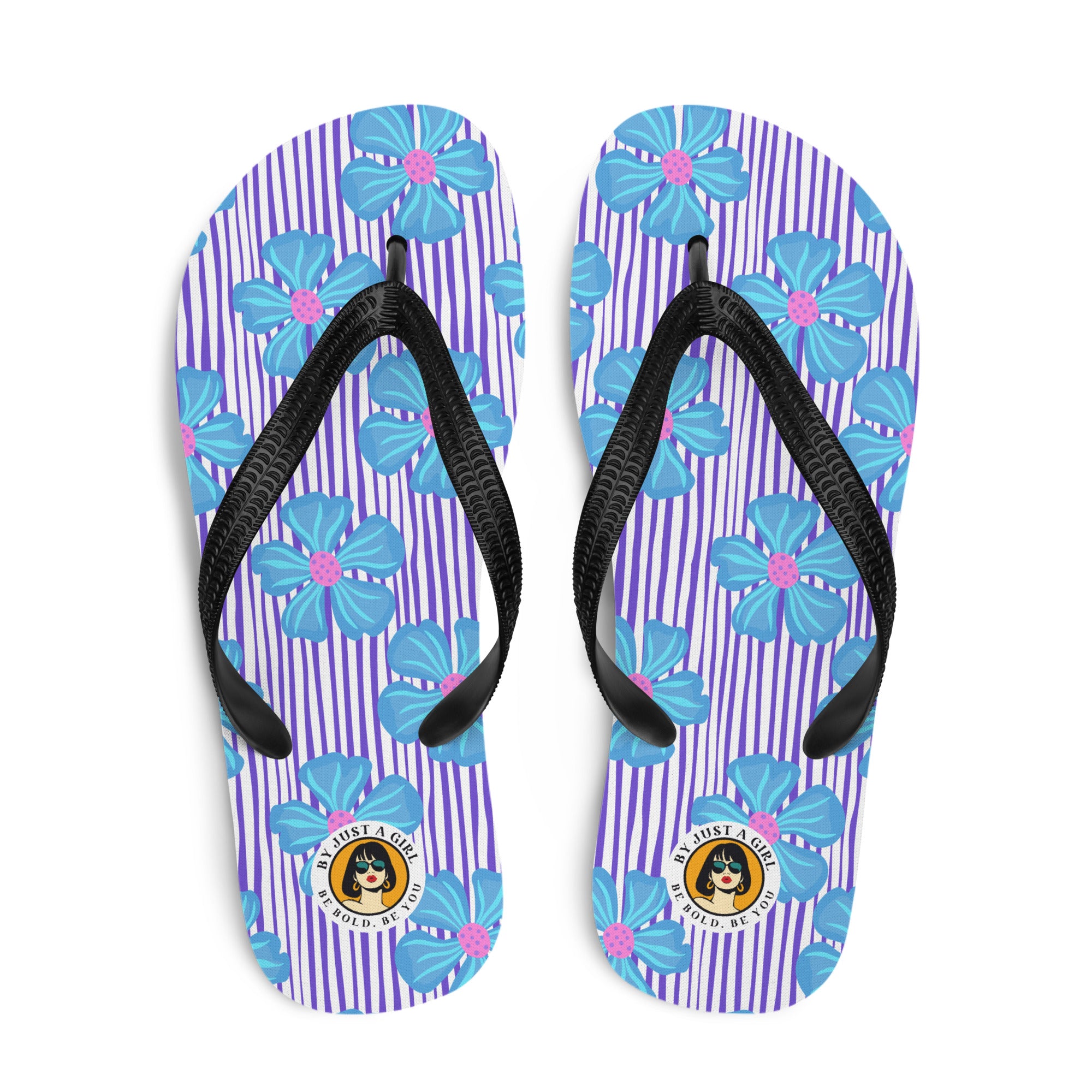 Ein Paar Flip-Flops mit einem Obermaterial, das ein Muster aus vertikalen lila und weißen Streifen zeigt, überlagert von großen, stilisierten blauen Blüten mit pinkfarbenen Zentren. Die Riemen sind schwarz und haben eine strukturierte Oberfläche. Auf der Ferse jedes Flip-Flops befindet sich ein rundes gelbes Emblem mit dem Text "BY JUST A GIRL" und "BE BOLD. BE YOU" sowie dem stilisierten Kopf einer Frau mit Bob-Frisur und Sonnenbrille.