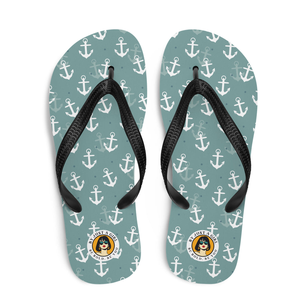 Ein Paar Flip-Flops mit einem Obermaterial in einem gedämpften Türkis- oder Blaugrünton, das mit einem wiederkehrenden Muster aus stilisierten weißen Ankern und kleinen grauen Punkten bedeckt ist. Die Riemen sind schwarz und haben eine strukturierte Oberfläche. Auf der Ferse jedes Flip-Flops befindet sich ein rundes gelbes Emblem mit dem Text "BY JUST A GIRL" und "BE BOLD. BE YOU" sowie dem stilisierten Kopf einer Frau mit Bob-Frisur und Sonnenbrille.