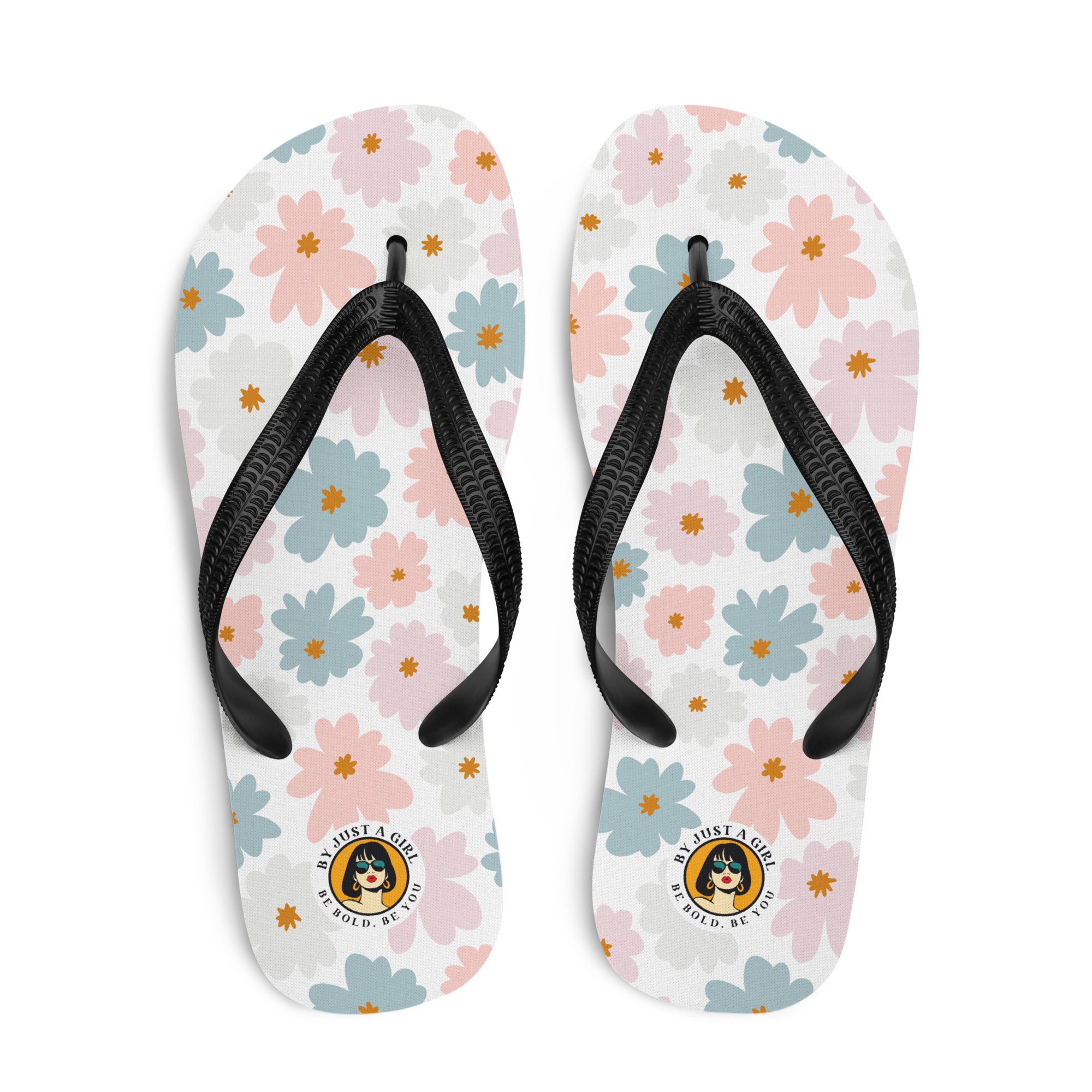 Ein Paar Flip-Flops mit einem weißen Obermaterial, das mit einem wiederkehrenden Muster aus stilisierten, fünfblättrigen Blüten in Pastellfarben wie Hellrosa, Hellblau und Beige bedeckt ist, ergänzt durch kleine orangefarbene Sterne in der Mitte einiger Blüten. Die Riemen sind schwarz und haben eine strukturierte Oberfläche. Auf der Ferse jedes Flip-Flops befindet sich ein rundes gelbes Emblem mit dem Text "BY JUST A GIRL" und "BE BOLD. BE YOU" sowie dem stilisierten Kopf einer Frau mit Bob-Frisur und Sonne