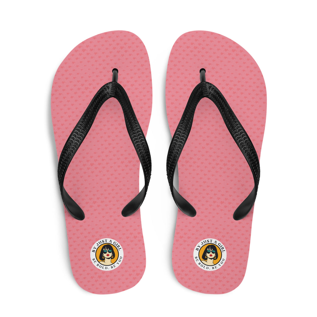 Ein Paar Flip-Flops mit einem Obermaterial, kleine Herzen in Rosa und Rot zeigt. Die Riemen sind schwarz und haben eine strukturierte Oberfläche. Auf der Ferse jedes Flip-Flops befindet sich ein rundes gelbes Emblem mit dem Text "BY JUST A GIRL" und "BE BOLD. BE YOU" sowie dem stilisierten Kopf einer Frau mit Bob-Frisur und Sonnenbrille.