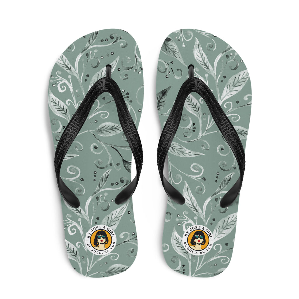Ein Paar Flip-Flops mit einem Obermaterial in einem gedämpften Salbeigrün oder Grau-Grün. Das Muster besteht aus stilisierten weißen und hellgrauen Blättern, Ranken und kleinen Punkten, die organisch über die Oberfläche verteilt sind. Die Riemen sind schwarz und haben eine strukturierte Oberfläche. Auf der Ferse jedes Flip-Flops befindet sich ein rundes gelbes Emblem mit dem Text "BY JUST A GIRL" und "BE BOLD. BE YOU" sowie dem stilisierten Kopf einer Frau mit Bob-Frisur und Sonnenbrille.