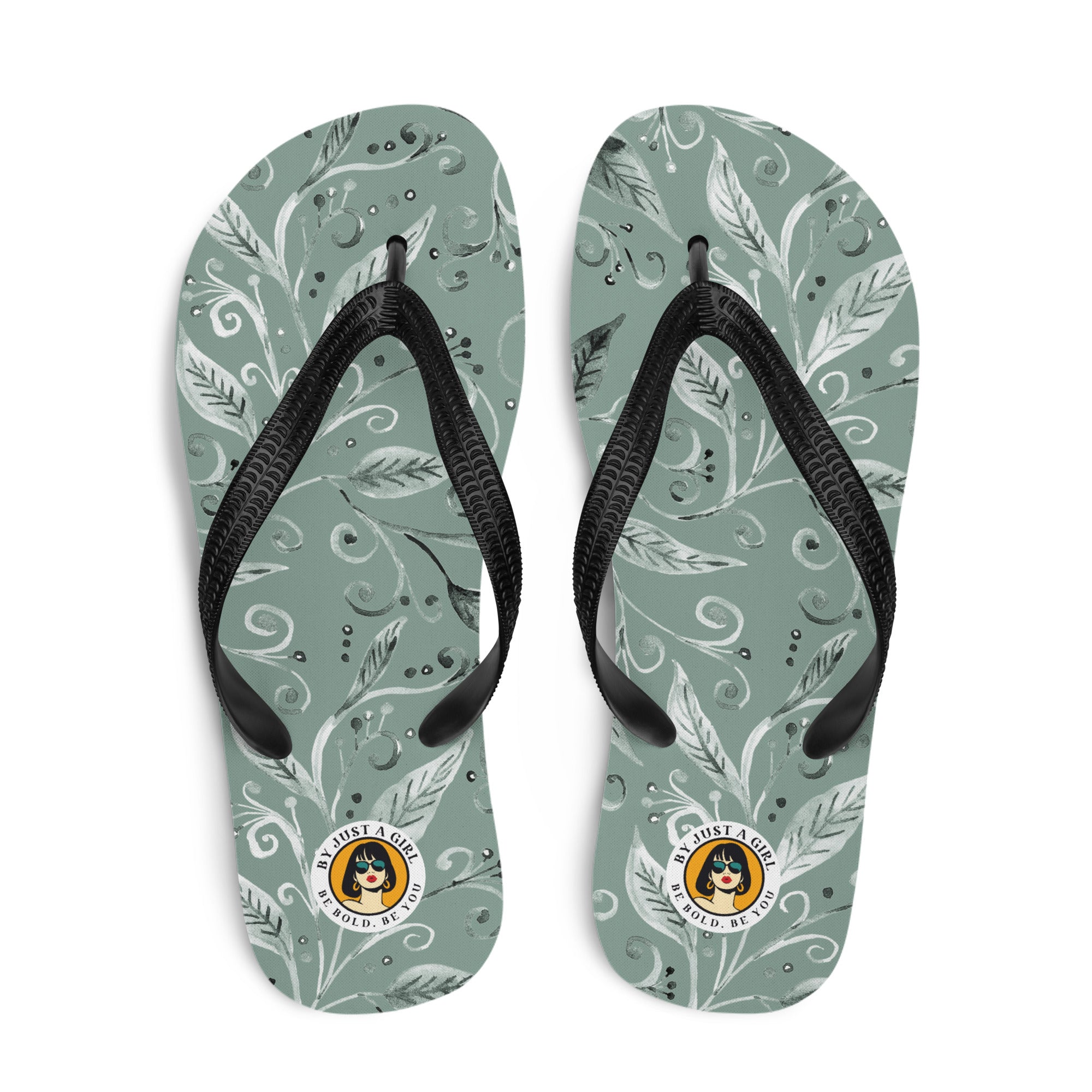 Ein Paar Flip-Flops mit einem Obermaterial in einem gedämpften Salbeigrün oder Grau-Grün. Das Muster besteht aus stilisierten weißen und hellgrauen Blättern, Ranken und kleinen Punkten, die organisch über die Oberfläche verteilt sind. Die Riemen sind schwarz und haben eine strukturierte Oberfläche. Auf der Ferse jedes Flip-Flops befindet sich ein rundes gelbes Emblem mit dem Text "BY JUST A GIRL" und "BE BOLD. BE YOU" sowie dem stilisierten Kopf einer Frau mit Bob-Frisur und Sonnenbrille.