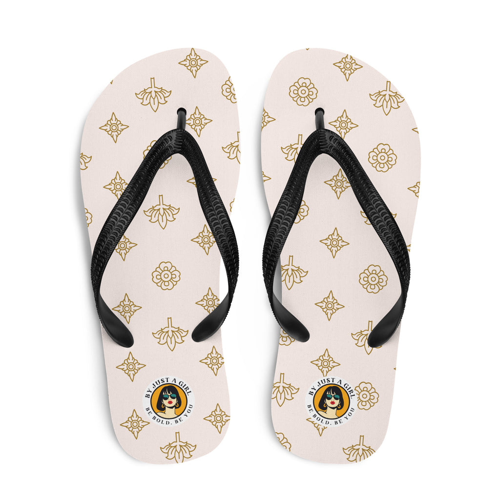 Ein Paar Flip-Flops mit einem hellbeigen oder cremefarbenen Obermaterial, das mit einem wiederkehrenden Muster aus stilisierten goldenen Lotusblüten und anderen floralen oder blattartigen Ornamenten bedeckt ist. Die Riemen sind schwarz und haben eine strukturierte Oberfläche. Auf der Ferse jedes Flip-Flops befindet sich ein rundes gelbes Emblem mit dem Text "BY JUST A GIRL" und "BE BOLD. BE YOU" sowie dem stilisierten Kopf einer Frau mit Bob-Frisur und Sonnenbrille.