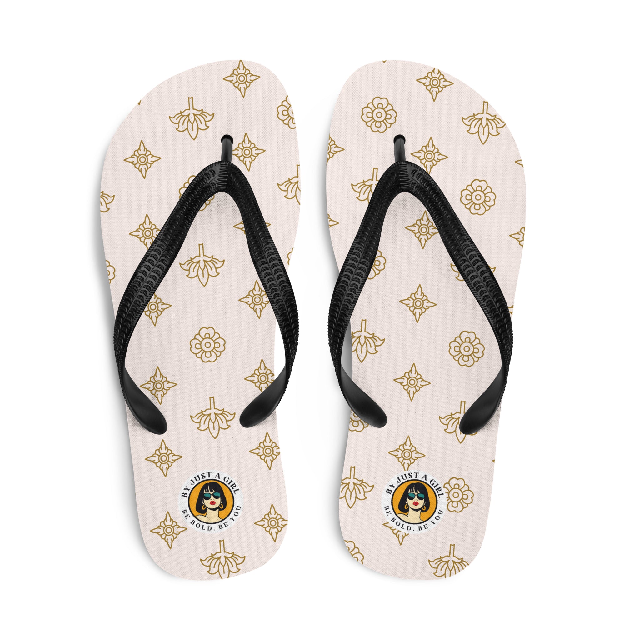 Ein Paar Flip-Flops mit einem hellbeigen oder cremefarbenen Obermaterial, das mit einem wiederkehrenden Muster aus stilisierten goldenen Lotusblüten und anderen floralen oder blattartigen Ornamenten bedeckt ist. Die Riemen sind schwarz und haben eine strukturierte Oberfläche. Auf der Ferse jedes Flip-Flops befindet sich ein rundes gelbes Emblem mit dem Text "BY JUST A GIRL" und "BE BOLD. BE YOU" sowie dem stilisierten Kopf einer Frau mit Bob-Frisur und Sonnenbrille.