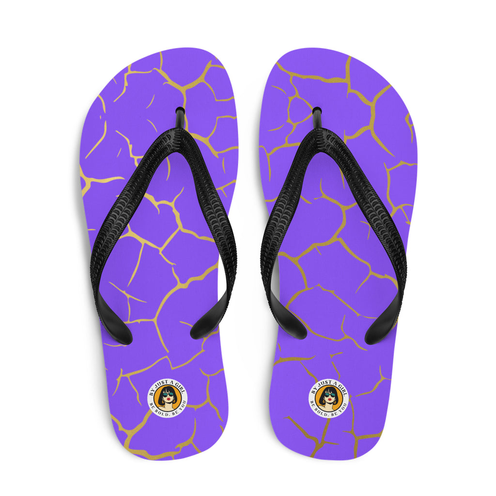Ein Paar Flip-Flops mit einem Obermaterial in einem kräftigen Violett, das von einem unregelmäßigen, spinnennetzartigen Muster aus goldenen Linien durchzogen ist, die wie Risse aussehen. Die Riemen sind schwarz und haben eine strukturierte Oberfläche. Auf der Ferse jedes Flip-Flops befindet sich ein rundes gelbes Emblem mit dem Text "BY JUST A GIRL" und "BE BOLD. BE YOU" sowie dem stilisierten Kopf einer Frau mit Bob-Frisur und Sonnenbrille.