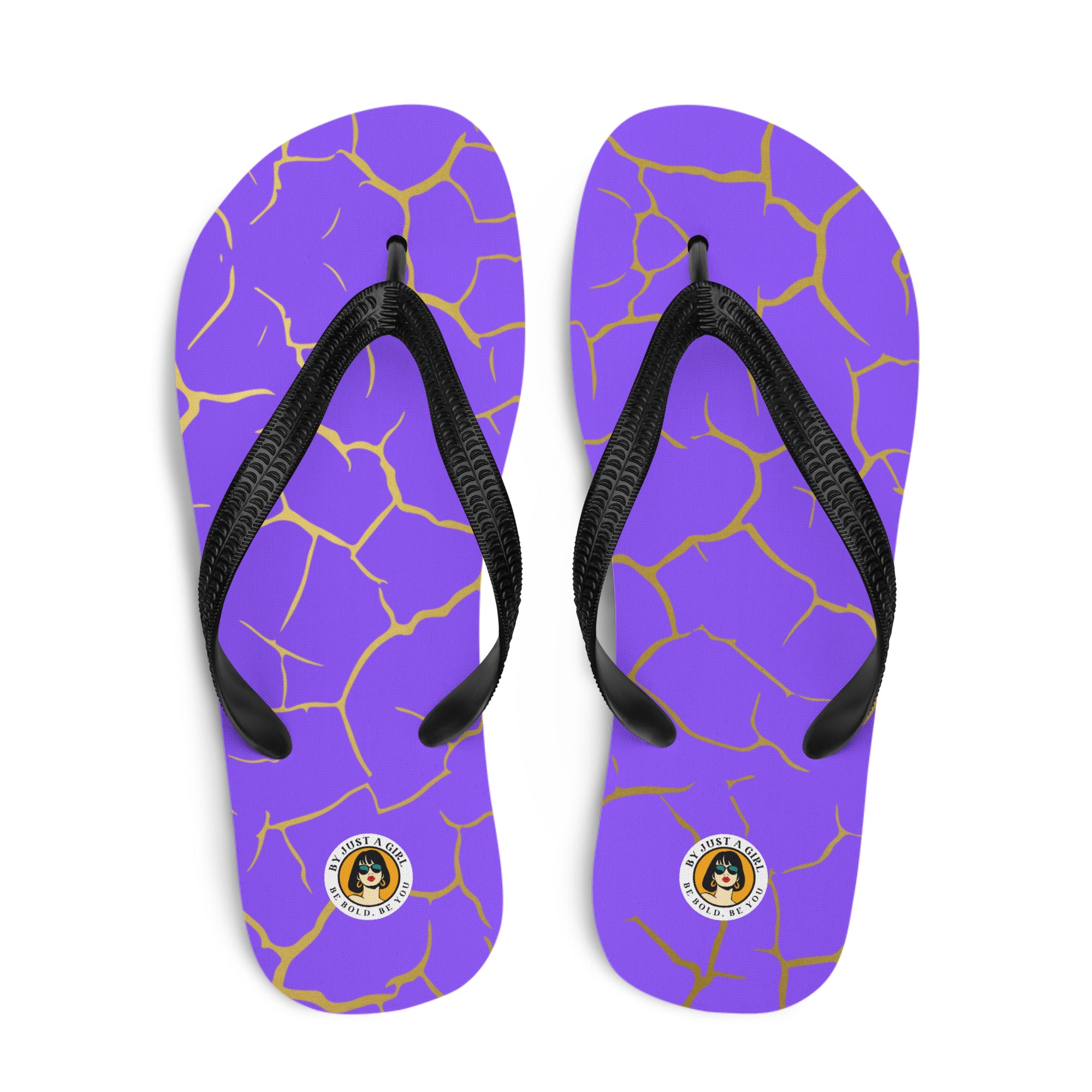 Ein Paar Flip-Flops mit einem Obermaterial in einem kräftigen Violett, das von einem unregelmäßigen, spinnennetzartigen Muster aus goldenen Linien durchzogen ist, die wie Risse aussehen. Die Riemen sind schwarz und haben eine strukturierte Oberfläche. Auf der Ferse jedes Flip-Flops befindet sich ein rundes gelbes Emblem mit dem Text "BY JUST A GIRL" und "BE BOLD. BE YOU" sowie dem stilisierten Kopf einer Frau mit Bob-Frisur und Sonnenbrille.