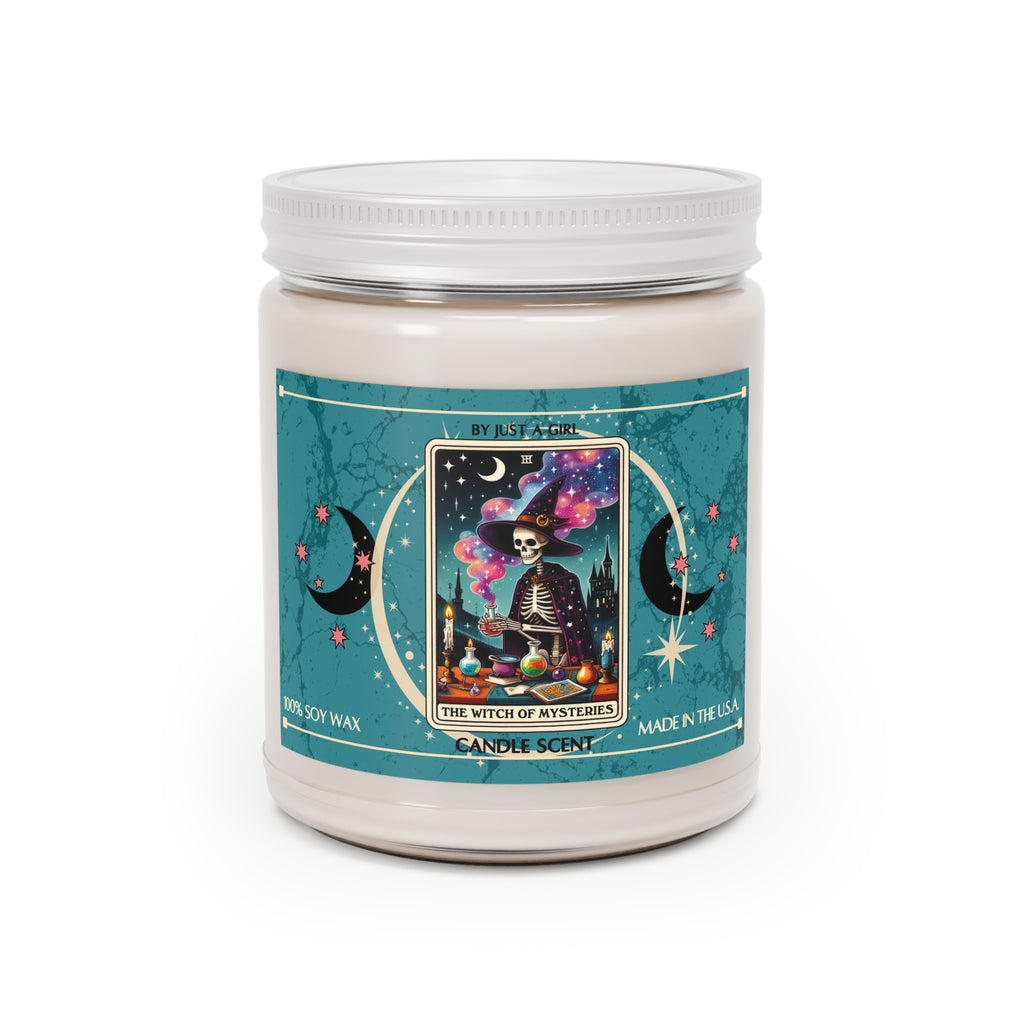 Eine weiße Kerze in einem durchsichtigen Glasgefäß mit weißem Deckel. Das Etikett ist türkisblau mit Mond- und Sternmotiven und zeigt in der Mitte eine goldgerahmte Tarotkarte mit einem Skelett in einem Hexenhut und Umhang, das an einem Tisch voller Zaubertränke und Bücher zaubert. Im Hintergrund ist eine dunkle Burg unter einem sternenklaren Nachthimmel zu sehen. Der Text auf dem Etikett lautet "BY JUST A GIRL", "III", "THE WITCH OF MYSTERIES" und "CANDLE SCENT". Unten steht "100% SOY WAX"
