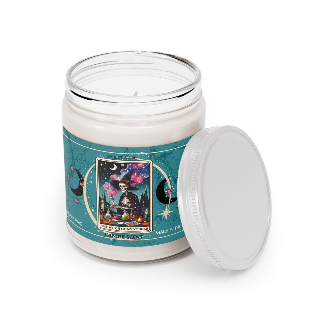 Eine weiße Kerze in einem durchsichtigen Glasgefäß mit weißem Deckel. Das Etikett ist türkisblau mit Mond- und Sternmotiven und zeigt in der Mitte eine goldgerahmte Tarotkarte mit einem Skelett in einem Hexenhut und Umhang, das an einem Tisch voller Zaubertränke und Bücher zaubert. Im Hintergrund ist eine dunkle Burg unter einem sternenklaren Nachthimmel zu sehen. Der Text auf dem Etikett lautet "BY JUST A GIRL", "III", "THE WITCH OF MYSTERIES" und "CANDLE SCENT". Unten steht "100% SOY WAX"