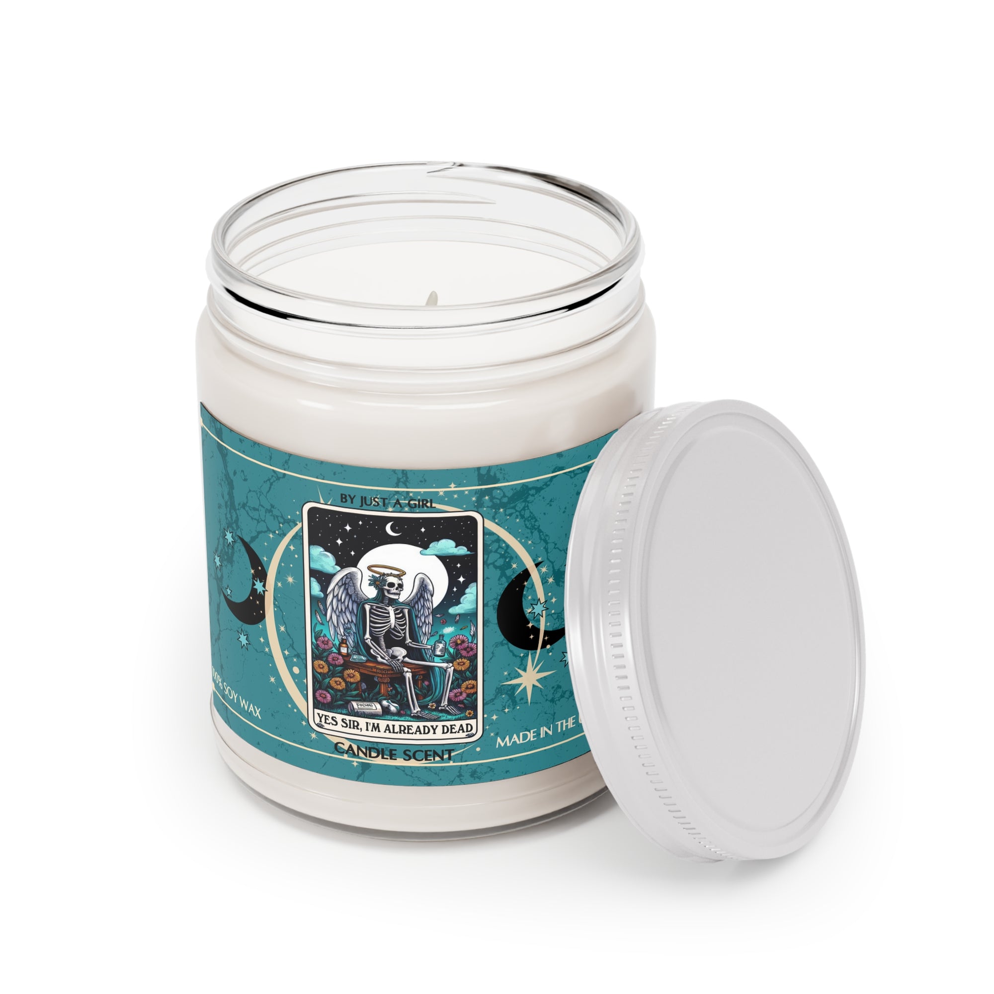 Eine weiße Kerze in einem durchsichtigen Glasgefäß mit weißem Deckel. Das Etikett ist türkisblau mit Mond- und Sternmotiven und zeigt in der Mitte eine goldgerahmte Illustration. Darauf ist ein geflügeltes Skelett zu sehen, das an einem Schreibtisch sitzt und schreibt, umgeben von Blumen und unter einem Mond. Der Text auf dem Etikett lautet "BY JUST A GIRL", "YES SIR, I'M ALREADY DEAD" und "CANDLE SCENT". Unten steht "100% SOY WAX" und "MADE IN THE U.S.A.".