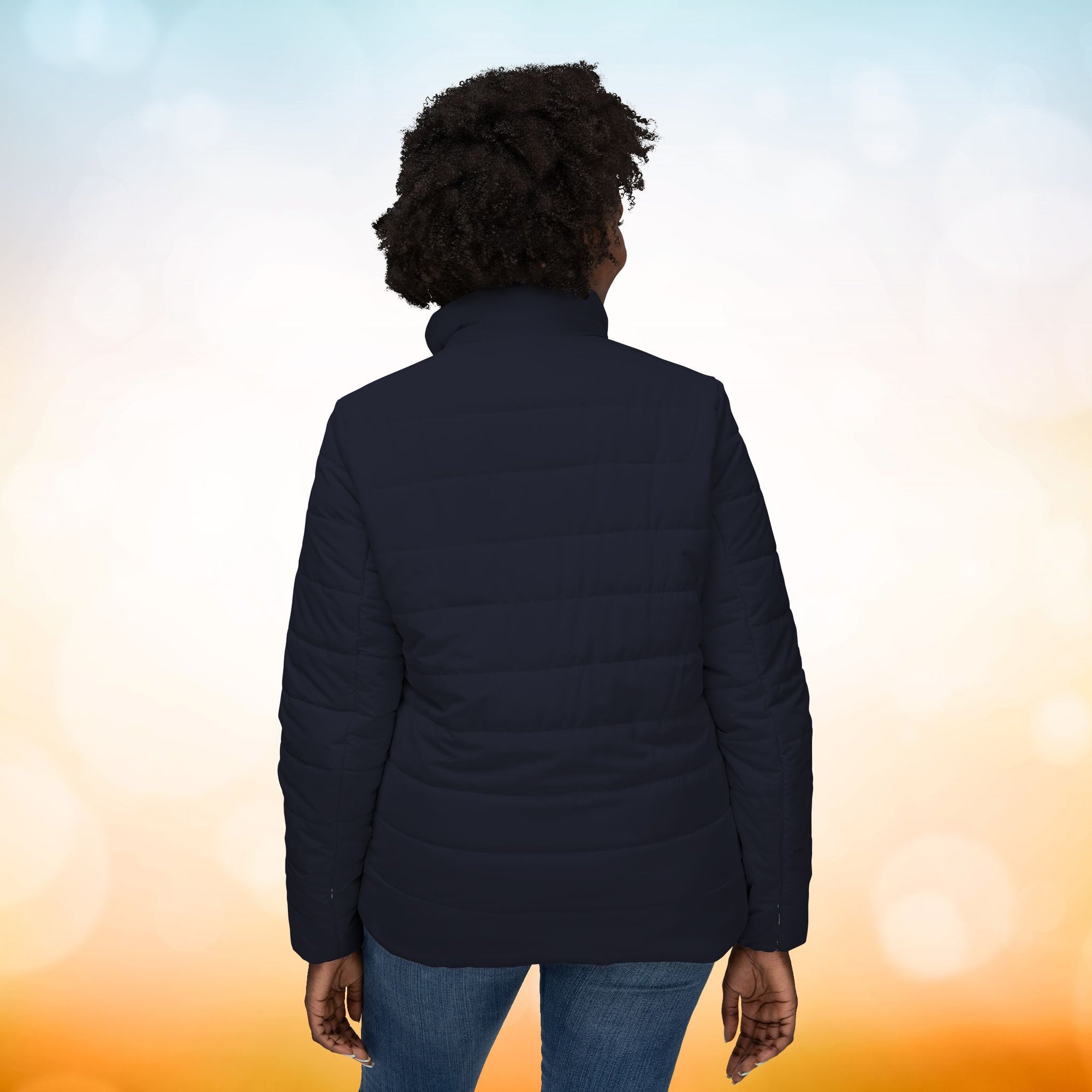 Das weibliche Model trägt eine Damen-Pufferjacke in einem Black Pearl Ton. Das Model präsentiert die Rückseite der Jacke. Die Jacke ist horizontal gesteppt, hat eine glatte Oberfläche sowie einen hohen Stehkragen. Designed, by Just a Girl