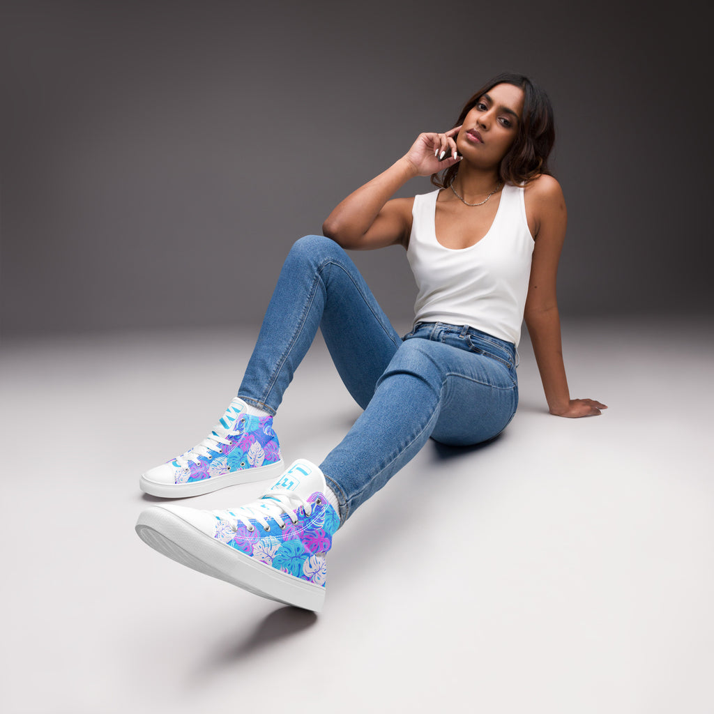 Ein weibliches Model trägt ein Paar High-Top-Canvas-Sneaker mit einem Muster aus tropischen Blättern in Blau, Lila und Pink. Die Sohlen, Zehenkappen und Schnürsenkel sind weiß. Die weißen Schuhzungen sind mit blauem Text "Leaf me" auf dem einen Schuh und "Wild" auf dem anderen beschriftet. Designed, by Just a Girl.