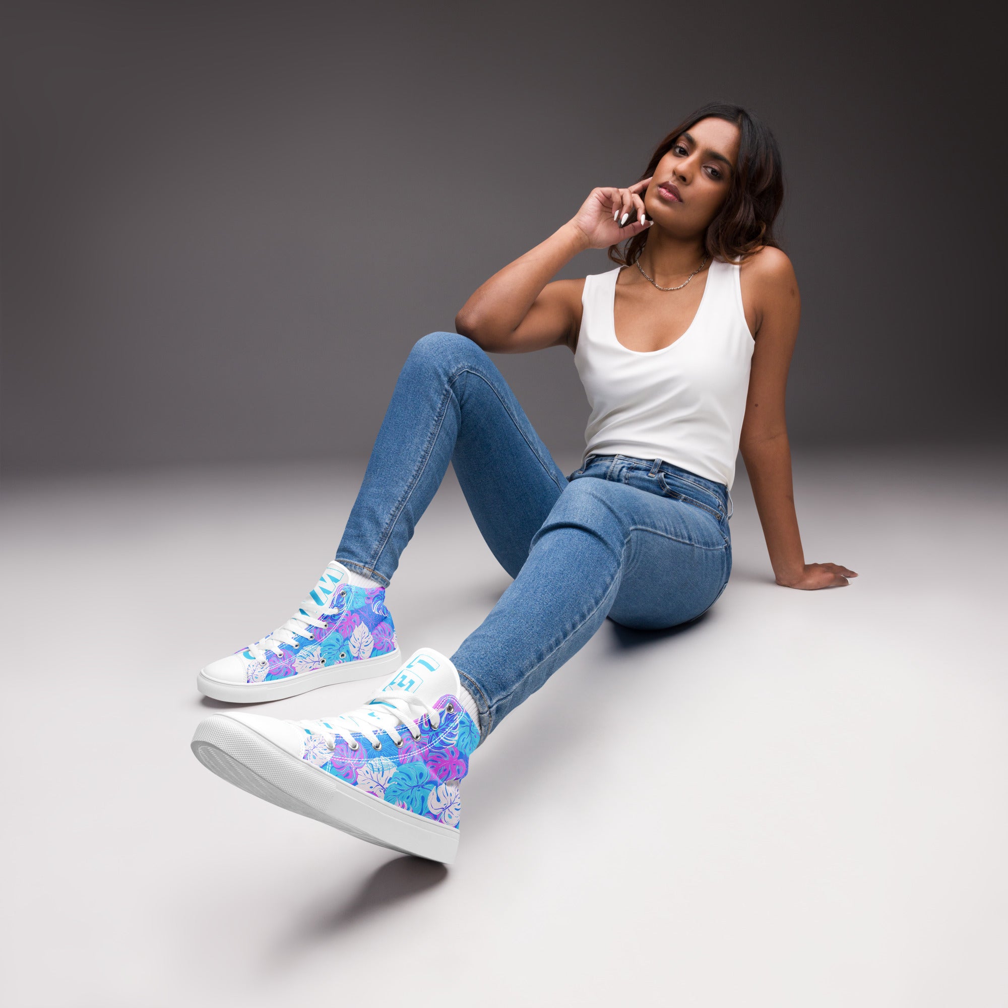 Ein weibliches Model trägt ein Paar High-Top-Canvas-Sneaker mit einem Muster aus tropischen Blättern in Blau, Lila und Pink. Die Sohlen, Zehenkappen und Schnürsenkel sind weiß. Die weißen Schuhzungen sind mit blauem Text "Leaf me" auf dem einen Schuh und "Wild" auf dem anderen beschriftet. Designed, by Just a Girl.