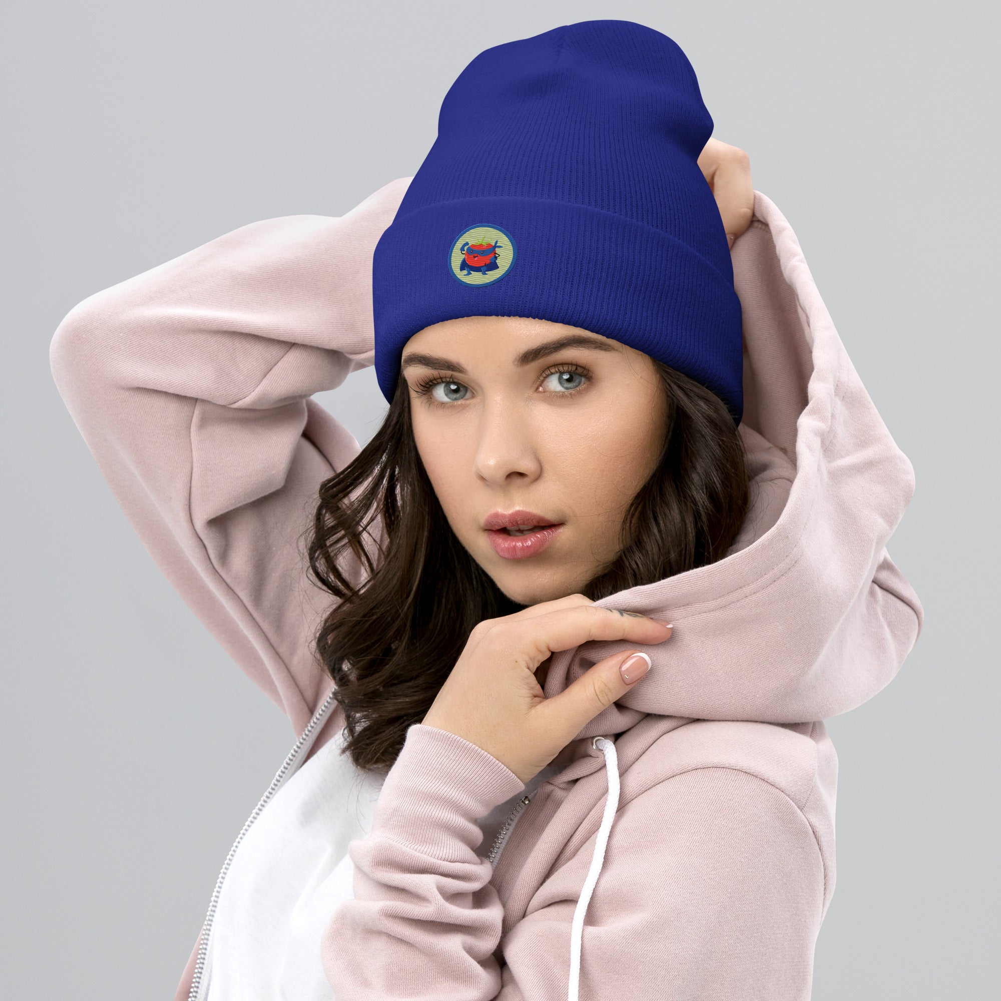 Das weibliche Model trägt ein Royal Blaues Cuffed Beanie mit aufgesticktem rotem Tomaten Superhelden Gesicht (Cartoon) in einem runden blauen Rahmen auf der Vorderseite. Designed, by Just a Girl.