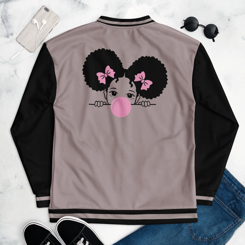 Eine Taupe farbene Bomberjacke mit schwarzen Ärmeln, Kragen und Saumbündchen. Auf der linken Brustseite ist eine Cartoon-Figur eines Mädchens mit dunklen Afro-Haaren, pinken Haarschleifen und einer großen rosa Kaugummi-Blase am Mund zu sehen. Die Jacke hat einen schwarzen Reißverschluss und schwarze Tascheneingriffe an der Vorderseite. Mockup Jacke liegend rückseite präsentiert mit Jeans, Schuhe & Sonnenbrille. By Just a Girl.