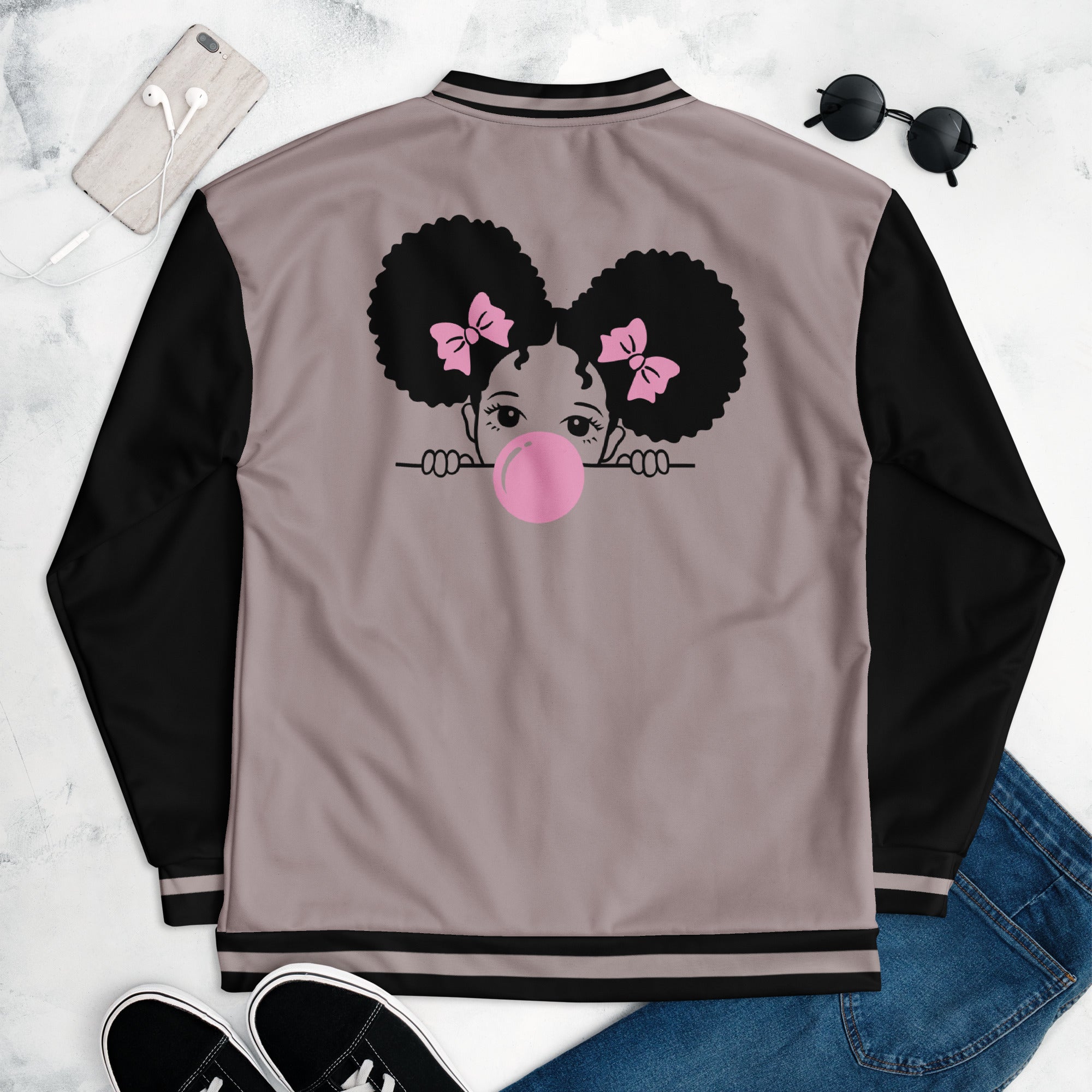 Eine Taupe farbene Bomberjacke mit schwarzen Ärmeln, Kragen und Saumbündchen. Auf der linken Brustseite ist eine Cartoon-Figur eines Mädchens mit dunklen Afro-Haaren, pinken Haarschleifen und einer großen rosa Kaugummi-Blase am Mund zu sehen. Die Jacke hat einen schwarzen Reißverschluss und schwarze Tascheneingriffe an der Vorderseite. Mockup Jacke liegend rückseite präsentiert mit Jeans, Schuhe & Sonnenbrille. By Just a Girl.