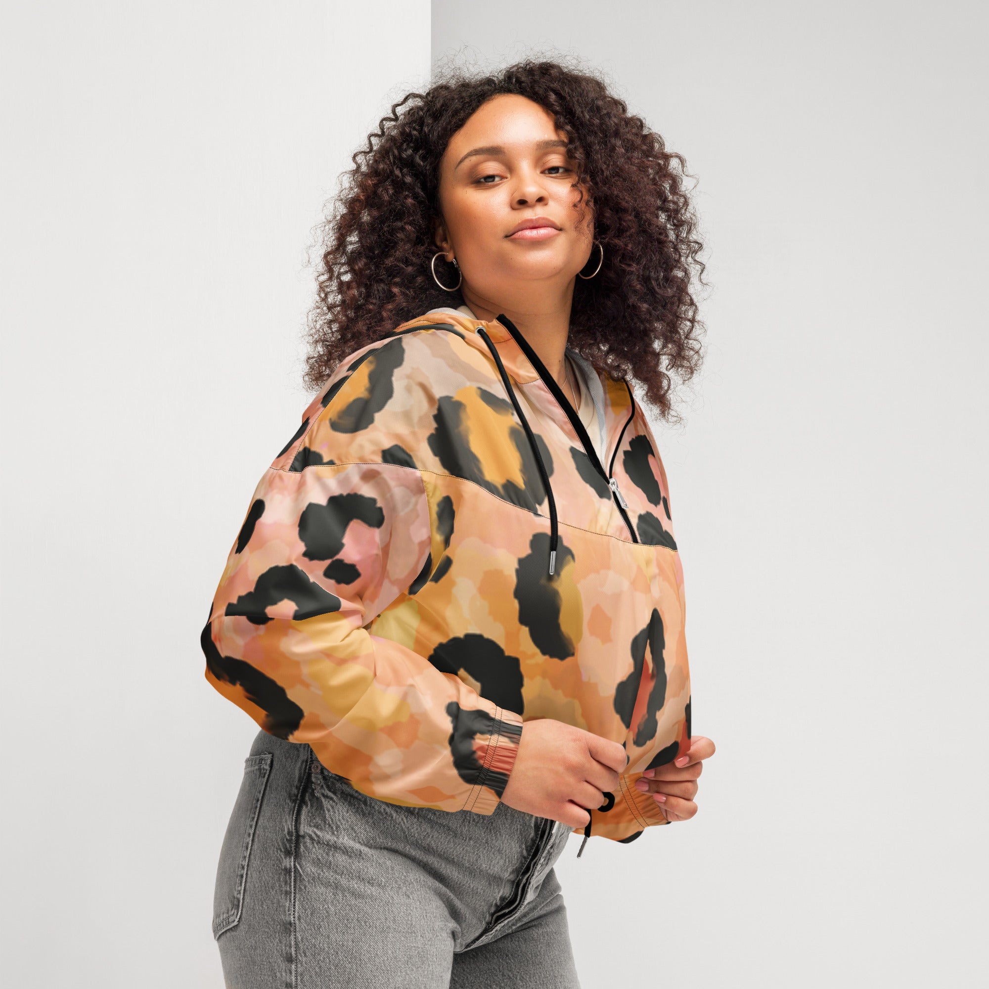Ein Damen Cropped Windbreaker mit einem abstrakten Leopardenmuster in Orange-, Rosa- und Schwarztönen. Die Jacke hat eine Kapuze, einen halben Reißverschluss mit schwarzen Kordeln und einen verstellbaren Saum. Mockup woman, seitliche Ansicht rechts. By Just a Girl.