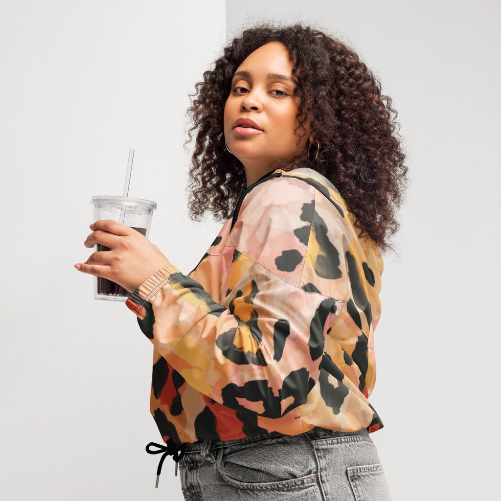 Ein Damen Cropped Windbreaker mit einem abstrakten Leopardenmuster in Orange-, Rosa- und Schwarztönen. Die Jacke hat eine Kapuze, einen halben Reißverschluss mit schwarzen Kordeln und einen verstellbaren Saum. Mockup woman seitliche Ansicht links. By Just a Girl.