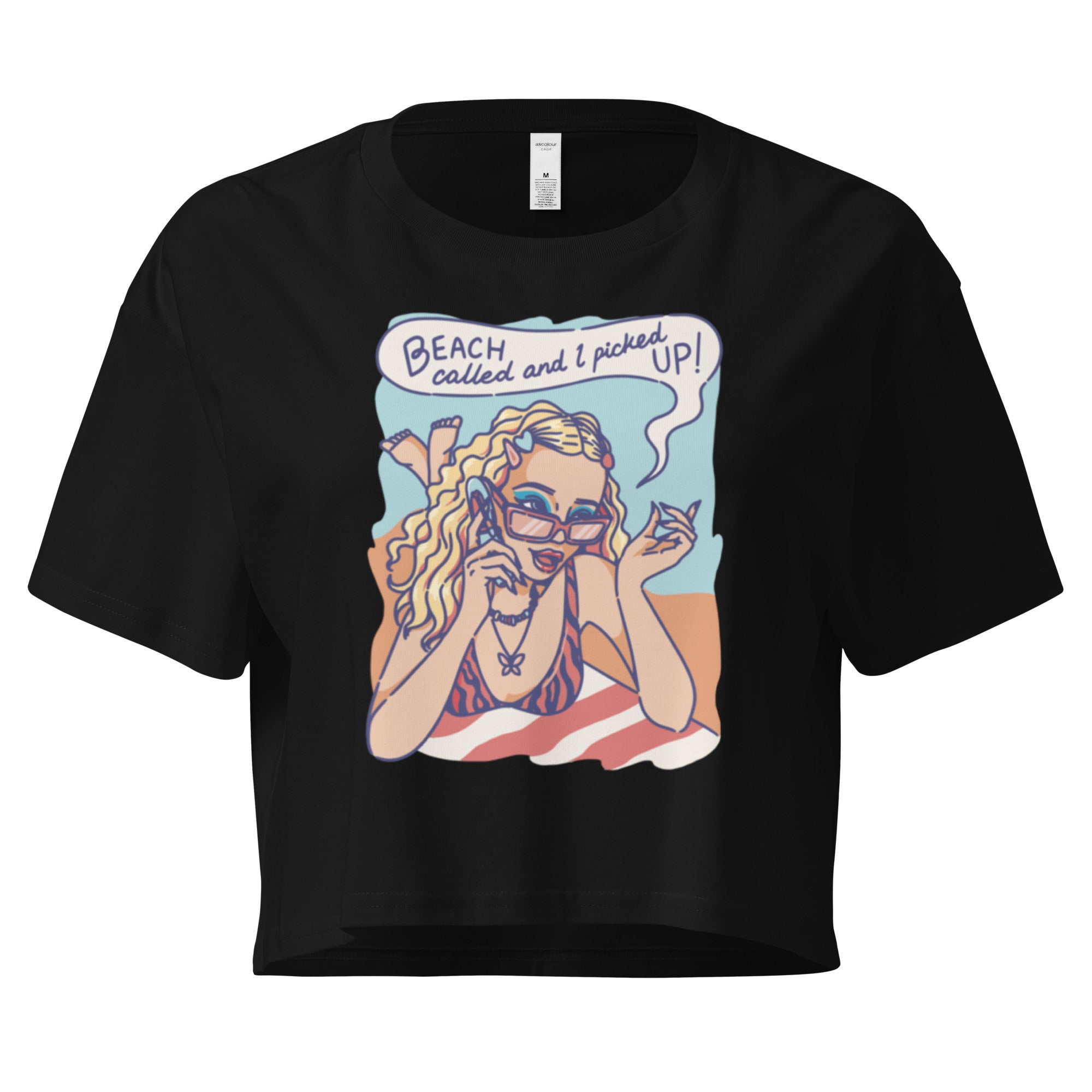 Ein schwarzes kurz geschnittenes Damen-T-Shirt (Crop Top) mit einer Retro-Illustration einer blonden Frau mit Sonnenbrille und gestreiftem Bikini, die entspannt am Strand liegt und telefoniert. Über ihr ist eine Sprechblase mit dem Text "BEACH called and I picked UP!" zu sehen. Der Hintergrund zeigt Sand und Meer.
