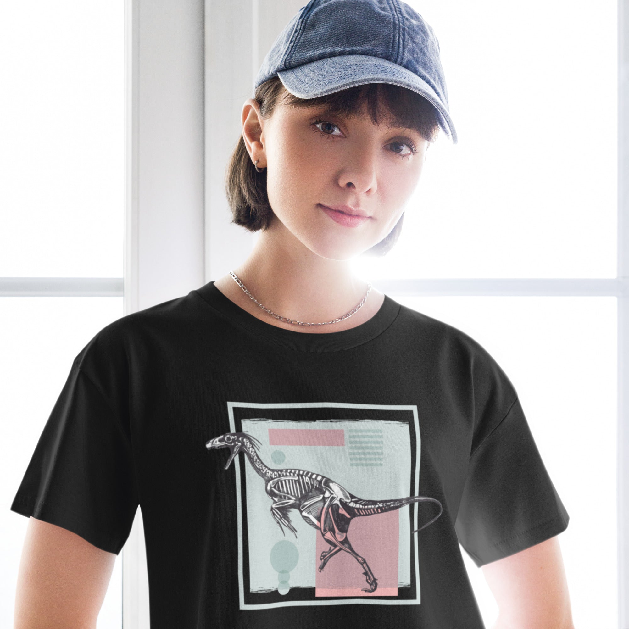 PEin schwarzes, kurz geschnittenes Damen-T-Shirt (Crop Top) mit einem weißen Kastenrahmen in der Mitte. Innerhalb des Rahmens befindet sich eine Illustration eines Dinosaurierskeletts, das auf einer Farbfeldfläche aus Rosa und Mintgrün platziert ist. Der Dinosaurier ist in einer Laufhaltung dargestellt.roduct mockup