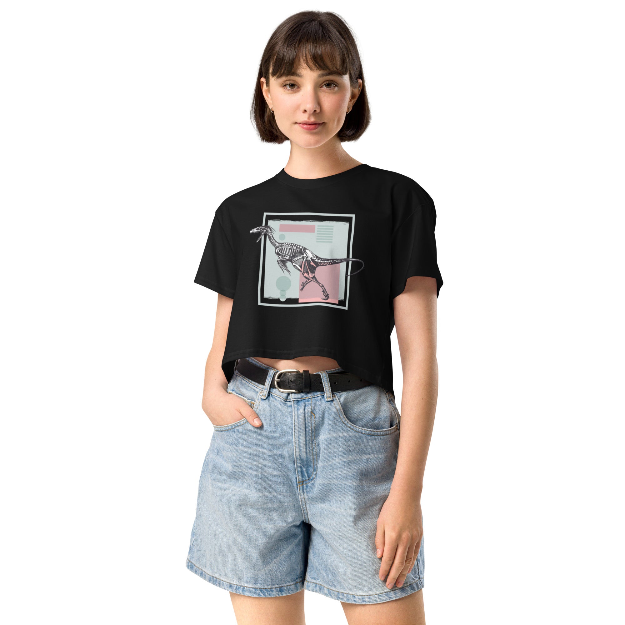 Ein schwarzes, kurz geschnittenes Damen-T-Shirt (Crop Top) mit einem weißen Kastenrahmen in der Mitte. Innerhalb des Rahmens befindet sich eine Illustration eines Dinosaurierskeletts, das auf einer Farbfeldfläche aus Rosa und Mintgrün platziert ist. Der Dinosaurier ist in einer Laufhaltung dargestellt.