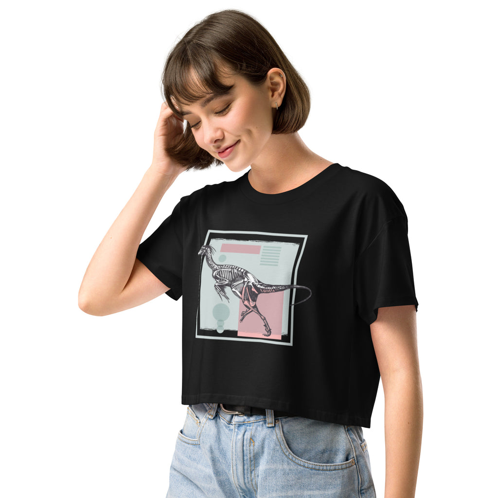 Ein schwarzes, kurz geschnittenes Damen-T-Shirt (Crop Top) mit einem weißen Kastenrahmen in der Mitte. Innerhalb des Rahmens befindet sich eine Illustration eines Dinosaurierskeletts, das auf einer Farbfeldfläche aus Rosa und Mintgrün platziert ist. Der Dinosaurier ist in einer Laufhaltung dargestellt.