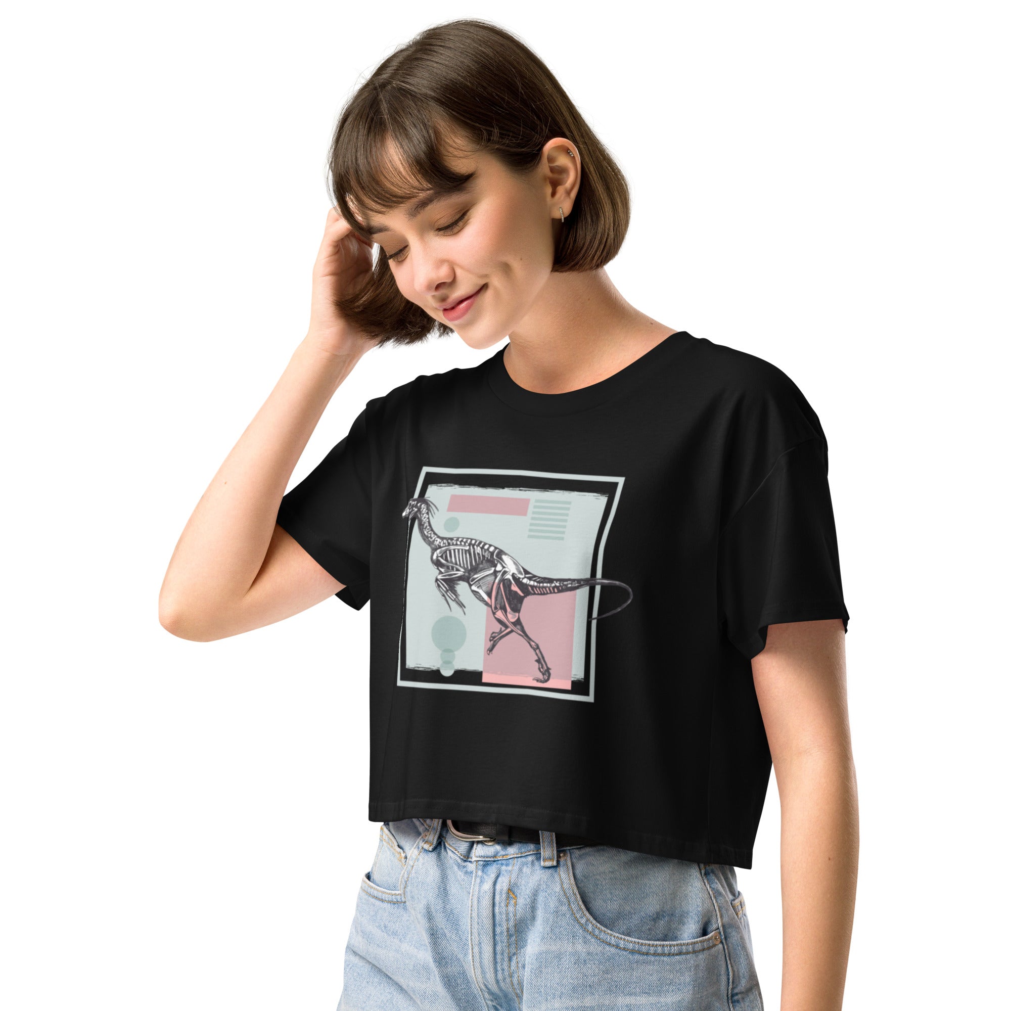 Ein schwarzes, kurz geschnittenes Damen-T-Shirt (Crop Top) mit einem weißen Kastenrahmen in der Mitte. Innerhalb des Rahmens befindet sich eine Illustration eines Dinosaurierskeletts, das auf einer Farbfeldfläche aus Rosa und Mintgrün platziert ist. Der Dinosaurier ist in einer Laufhaltung dargestellt.