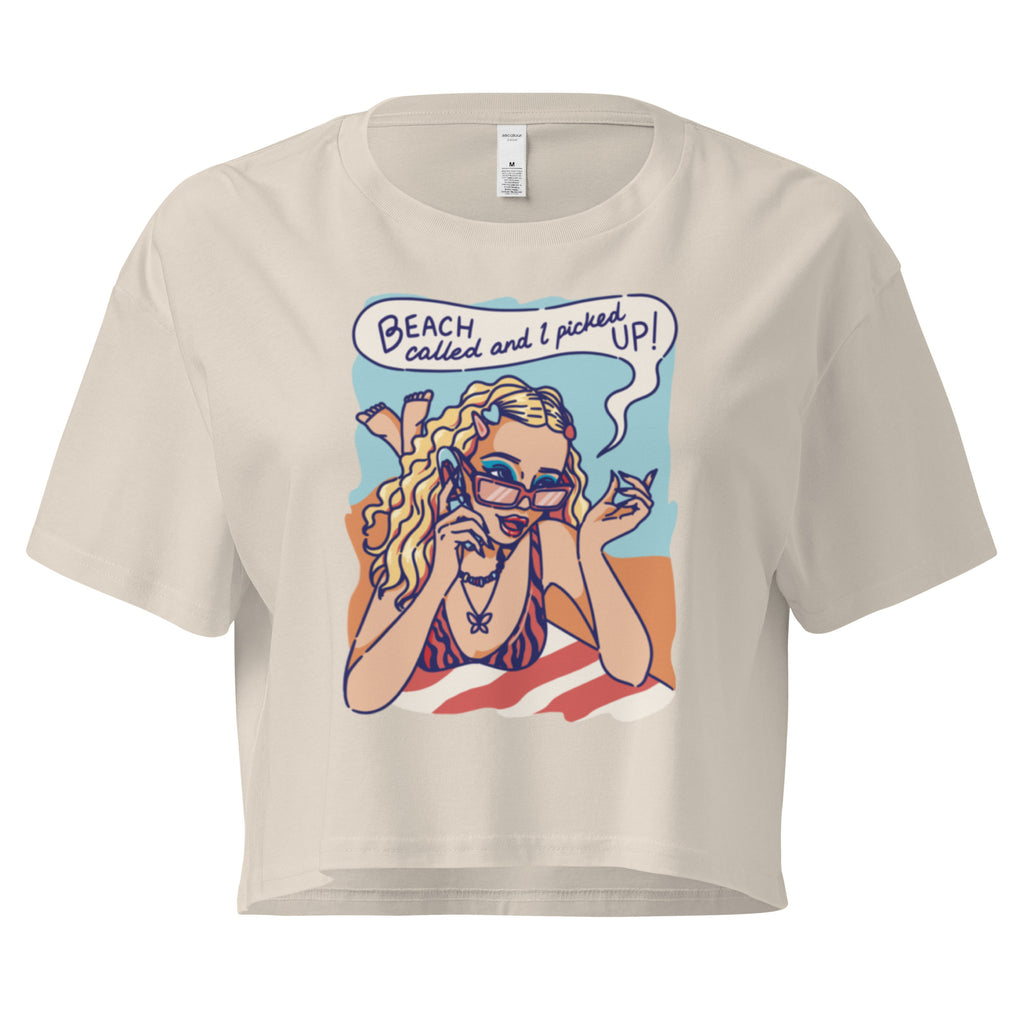 Ein bone farbenes kurz geschnittenes Damen-T-Shirt (Crop Top) mit einer Retro-Illustration einer blonden Frau mit Sonnenbrille und gestreiftem Bikini, die entspannt am Strand liegt und telefoniert. Über ihr ist eine Sprechblase mit dem Text "BEACH called and I picked UP!" zu sehen. Der Hintergrund zeigt Sand und Meer.