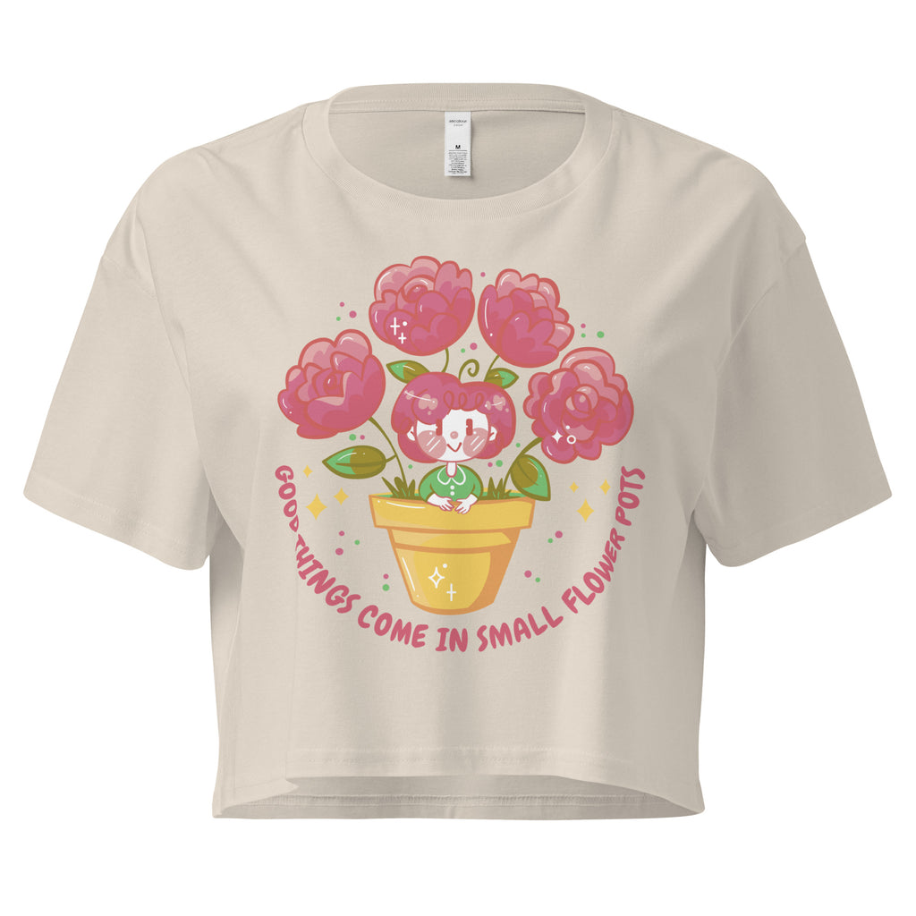 Ein bone farbenes kurz geschnittenes Damen-T-Shirt (Crop Top) mit einer Illustration einer kleinen, lächelnden Figur in einem gelben Blumentopf. Um den Blumentopf herum sind mehrere große, rosa Blüten und grüne Blätter. Der Text "GOOD THINGS COME IN SMALL FLOWER POTS" ist in einem Bogen um das Motiv herum angeordnet, mit kleinen Sternen und Punkten.