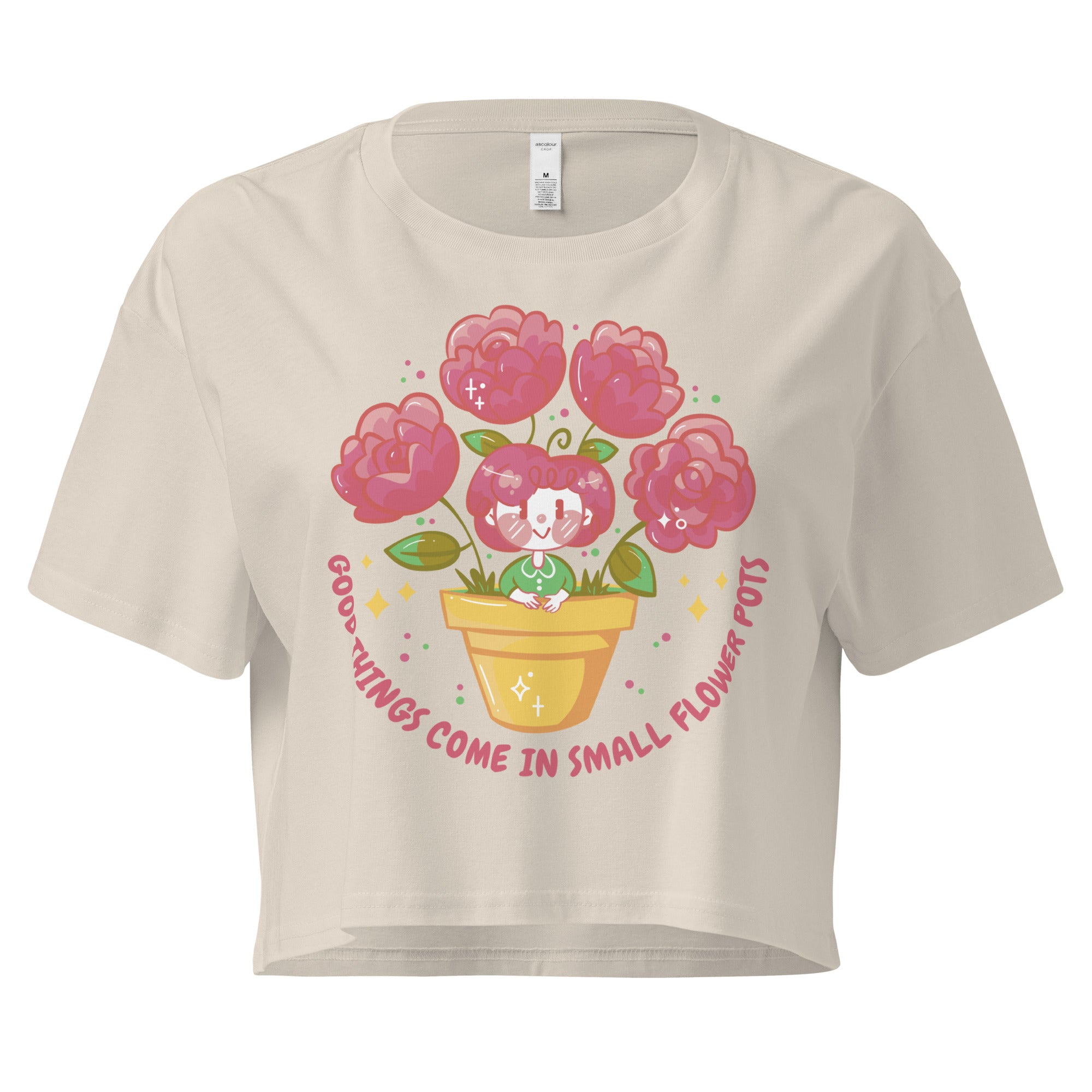 Ein bone farbenes kurz geschnittenes Damen-T-Shirt (Crop Top) mit einer Illustration einer kleinen, lächelnden Figur in einem gelben Blumentopf. Um den Blumentopf herum sind mehrere große, rosa Blüten und grüne Blätter. Der Text "GOOD THINGS COME IN SMALL FLOWER POTS" ist in einem Bogen um das Motiv herum angeordnet, mit kleinen Sternen und Punkten.