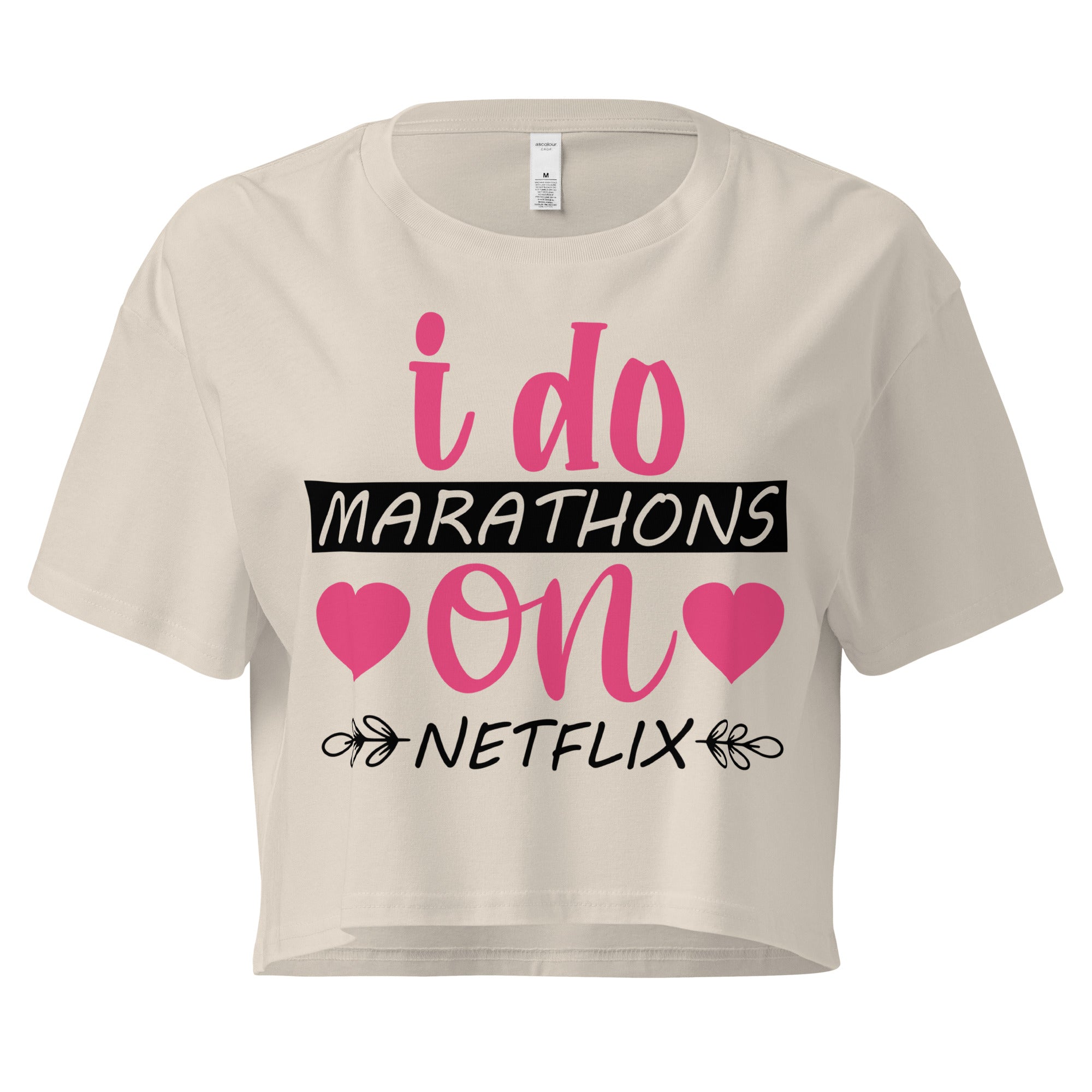 Ein bone oder Beige farbenes, kurz geschnittenes Damen-T-Shirt (Crop Top) mit dem Text "i do MARATHONS on NETFLIX" auf der Vorderseite. Der Text "i do" und "on" ist in Pink, "MARATHONS" in Schwarz auf einem schwarzen Balken, und "NETFLIX" ebenfalls in Schwarz, umgeben von kleinen schwarzen Herzen und floralen Ranken.