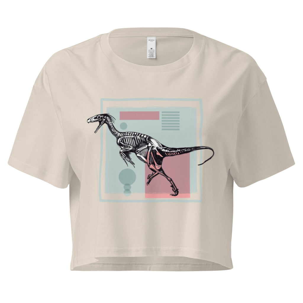 Ein beige farbenes, kurz geschnittenes Damen-T-Shirt (Crop Top) mit einem weißen Kastenrahmen in der Mitte. Innerhalb des Rahmens befindet sich eine Illustration eines Dinosaurierskeletts, das auf einer Farbfeldfläche aus Rosa und Mintgrün platziert ist. Der Dinosaurier ist in einer Laufhaltung dargestellt.