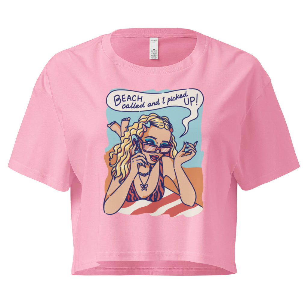 Ein bubblegum farbenes kurz geschnittenes Damen-T-Shirt (Crop Top) mit einer Retro-Illustration einer blonden Frau mit Sonnenbrille und gestreiftem Bikini, die entspannt am Strand liegt und telefoniert. Über ihr ist eine Sprechblase mit dem Text "BEACH called and I picked UP!" zu sehen. Der Hintergrund zeigt Sand und Meer.