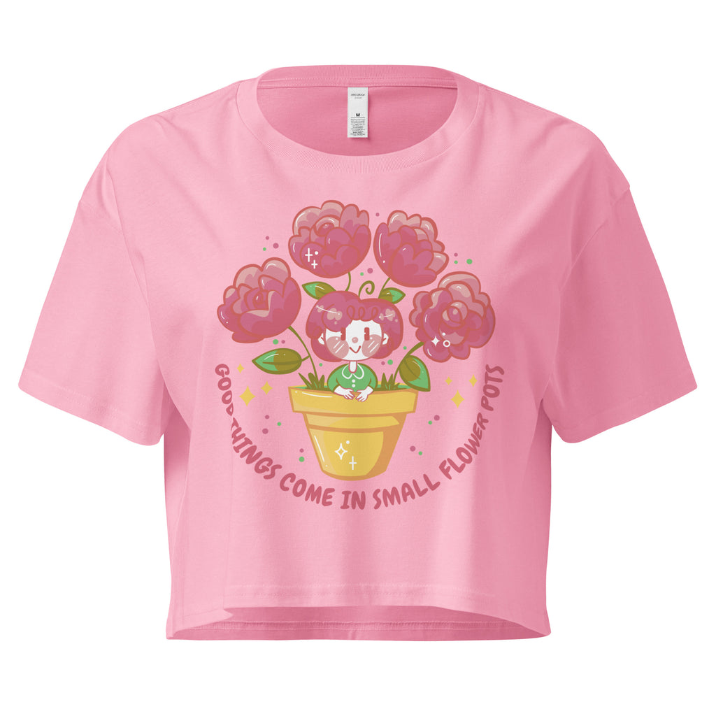 Ein bubblegum farbenes kurz geschnittenes Damen-T-Shirt (Crop Top) mit einer Illustration einer kleinen, lächelnden Figur in einem gelben Blumentopf. Um den Blumentopf herum sind mehrere große, rosa Blüten und grüne Blätter. Der Text "GOOD THINGS COME IN SMALL FLOWER POTS" ist in einem Bogen um das Motiv herum angeordnet, mit kleinen Sternen und Punkten.