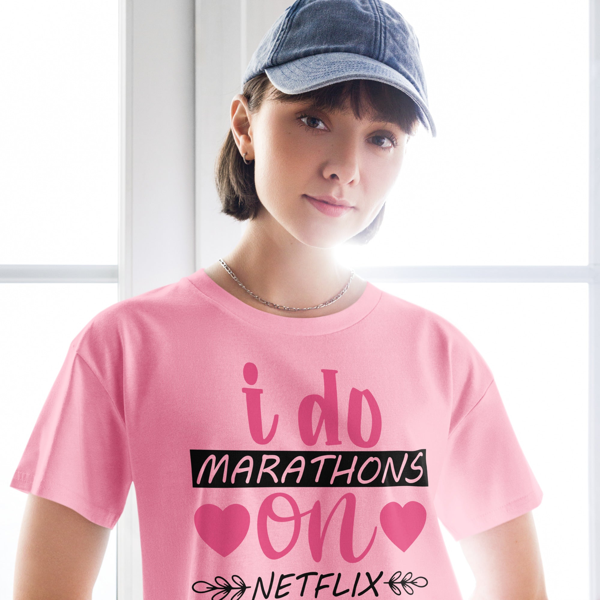 Ein "Bubblegum-Pink" farbenes, kurz geschnittenes Damen-T-Shirt (Crop Top) mit dem Text "i do MARATHONS on NETFLIX" auf der Vorderseite. Der Text "i do" und "on" ist in Pink, "MARATHONS" in Schwarz auf einem schwarzen Balken, und "NETFLIX" ebenfalls in Schwarz, umgeben von kleinen schwarzen Herzen und floralen Ranken.