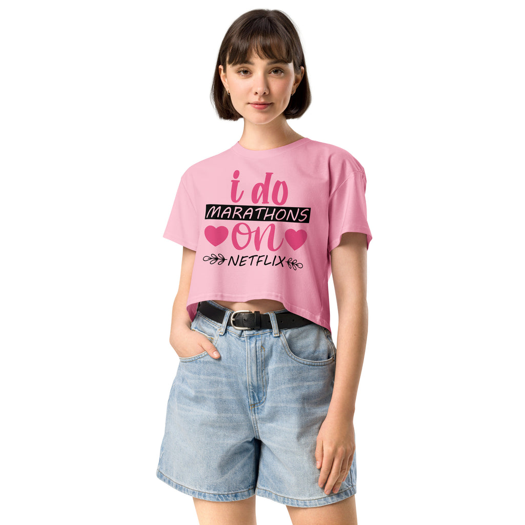 Ein "Bubblegum-Pink" farbenes, kurz geschnittenes Damen-T-Shirt (Crop Top) mit dem Text "i do MARATHONS on NETFLIX" auf der Vorderseite. Der Text "i do" und "on" ist in Pink, "MARATHONS" in Schwarz auf einem schwarzen Balken, und "NETFLIX" ebenfalls in Schwarz, umgeben von kleinen schwarzen Herzen und floralen Ranken.