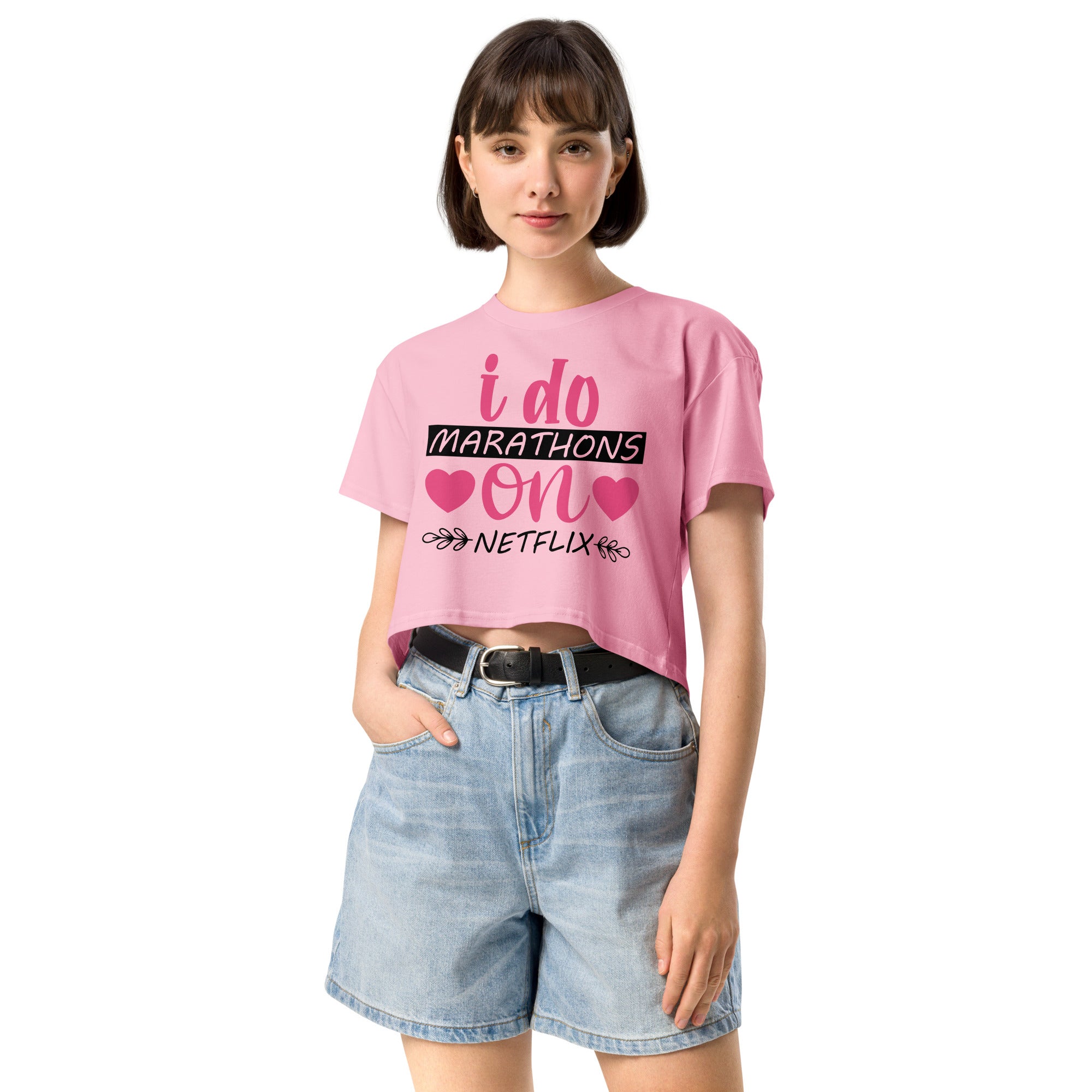 Ein "Bubblegum-Pink" farbenes, kurz geschnittenes Damen-T-Shirt (Crop Top) mit dem Text "i do MARATHONS on NETFLIX" auf der Vorderseite. Der Text "i do" und "on" ist in Pink, "MARATHONS" in Schwarz auf einem schwarzen Balken, und "NETFLIX" ebenfalls in Schwarz, umgeben von kleinen schwarzen Herzen und floralen Ranken.